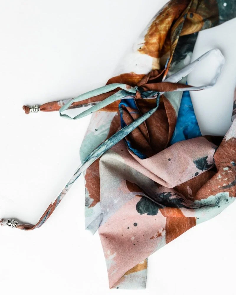 Colorful silk scarf with tie-dye pattern in shades of pink, blue, brown, and green, with silver decorative tips on the ends, resting on a white surface.