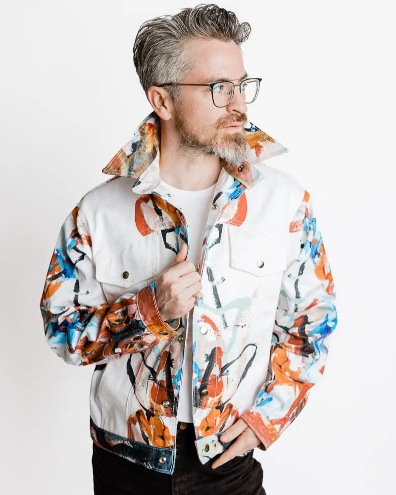 A man with gray hair and beard wearing glasses, a white jacket with colorful abstract patterns, and a white shirt, posing against a plain white background.