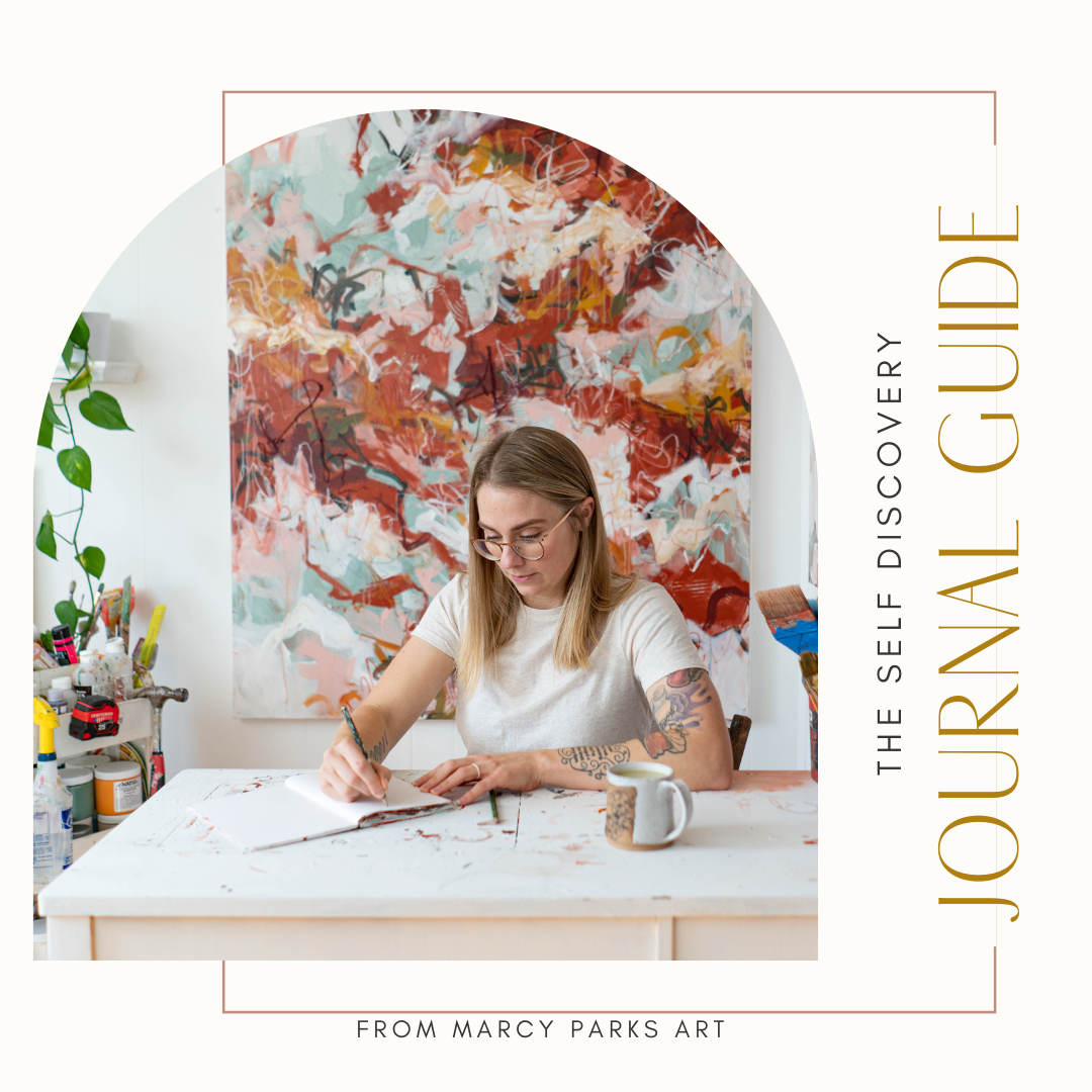 A woman with glasses drawing in a sketchbook at a table with art supplies, with an abstract painting in shades of red, pink, and blue hanging on the wall behind her.