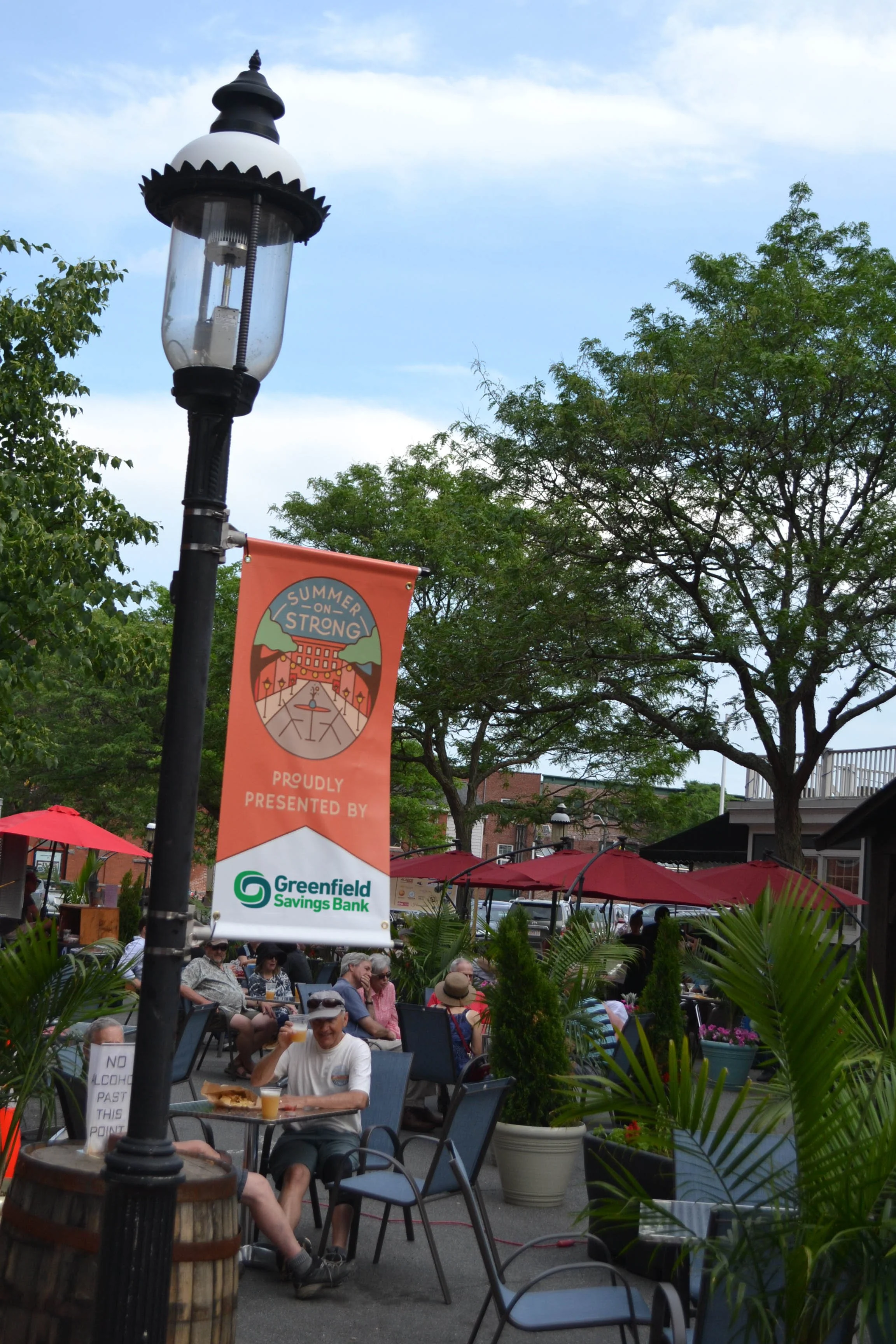 Summer on Strong at Strong Avenue in Northampton - people enjoying outdoor seating, with the Greenfield Savings Bank sponsorship banner in the foreground