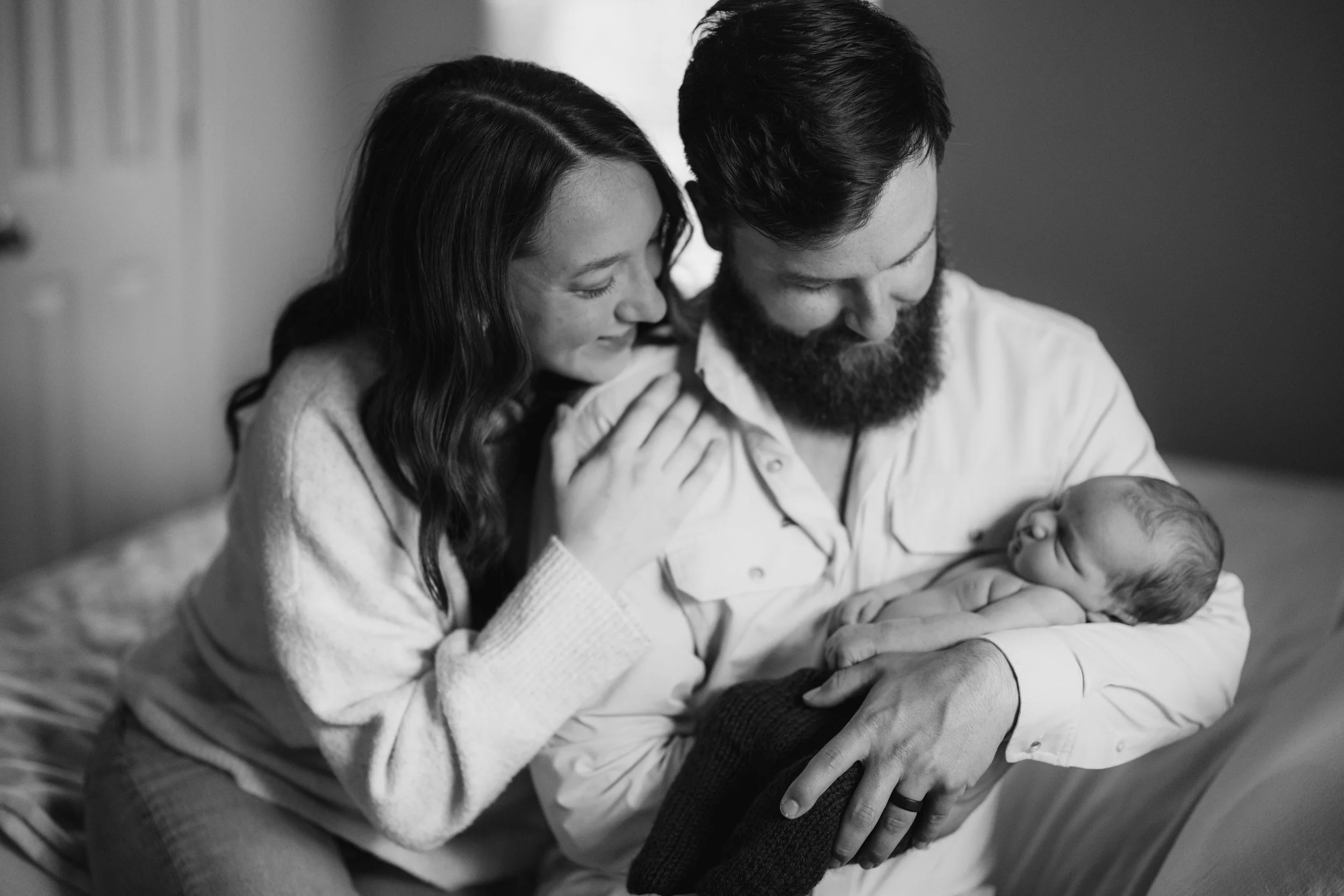 A black and white photo of a woman and a man holding a newborn baby, with their heads close together and eyes closed, in a tender moment in a cozy indoor setting.