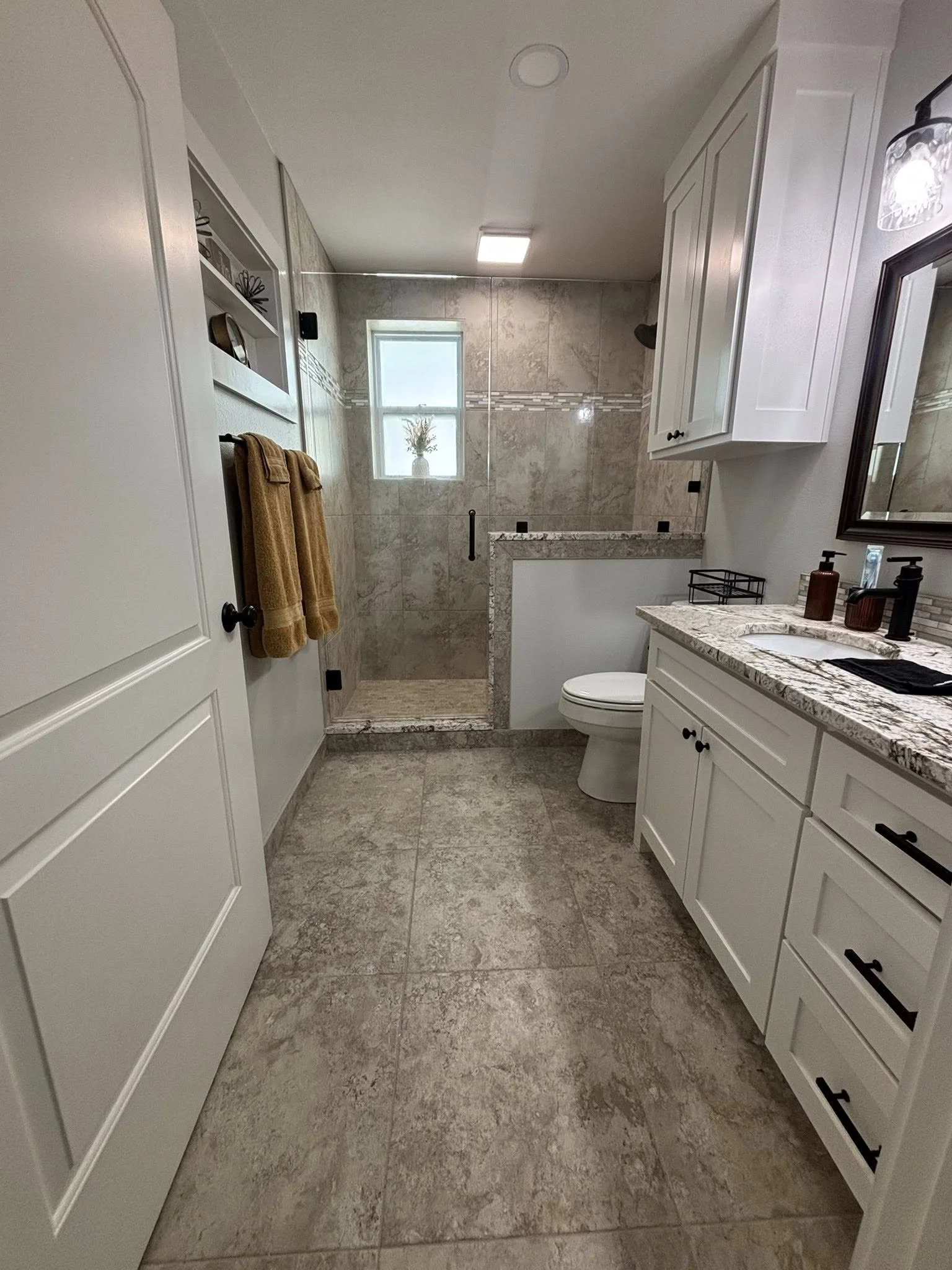 A modern bathroom with a granite countertop, white cabinets, a toilet, a glass-enclosed shower with beige tiles, a small window. ETX Tyler Texas Residential Interior bathroom remodel.