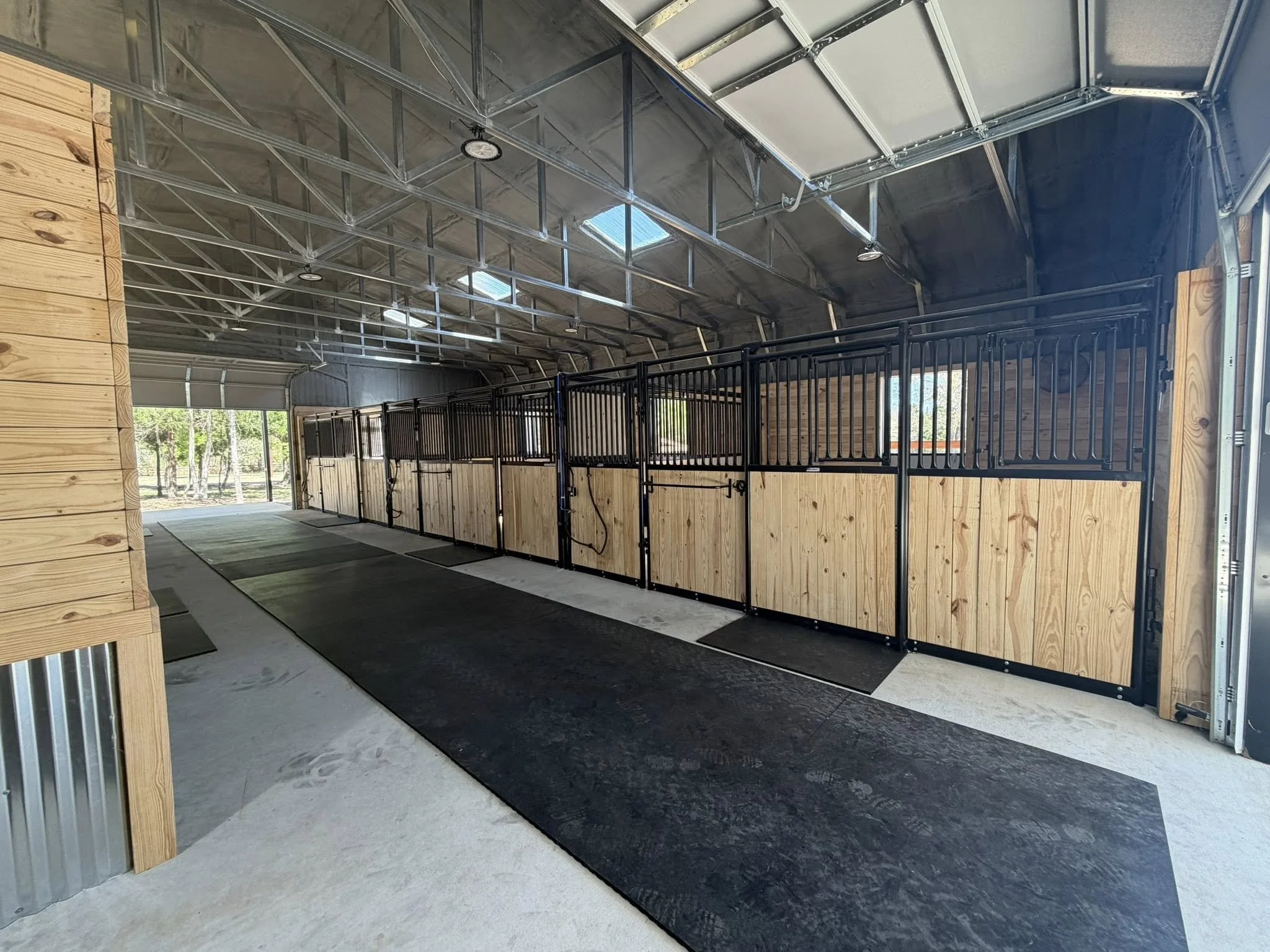 The interior of a horse barn with black metal stalls and light wood walls, a concrete floor, and overhead lighting. There are large garage doors open to the outside. ETX Tyler Texas Residential New Construction