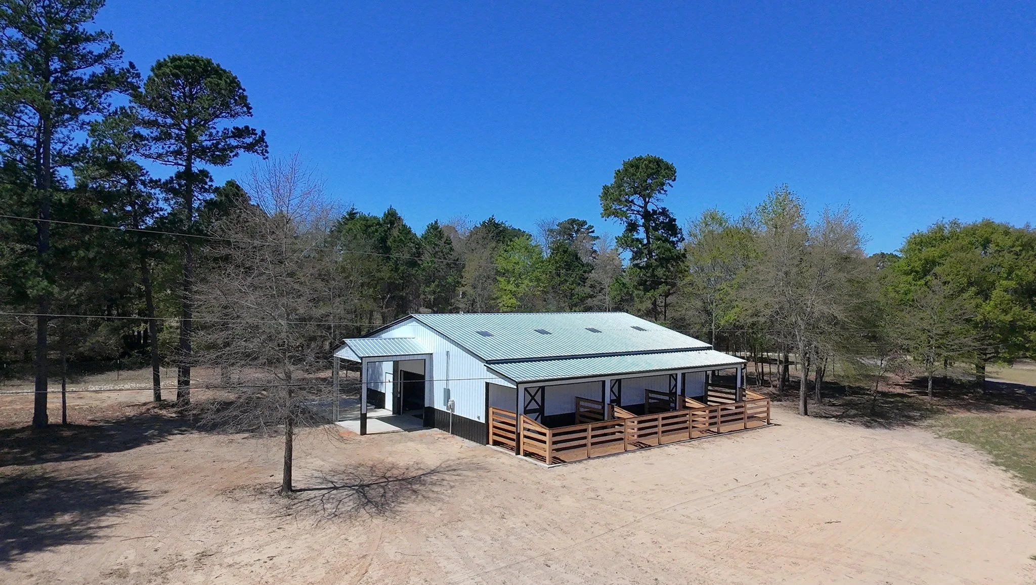 A white metal building with a covered porch, situated in a cleared area with dirt ground and a wooden fence, surrounded by tall trees and a bright blue sky. ETX Tyler Texas Residential New Construction