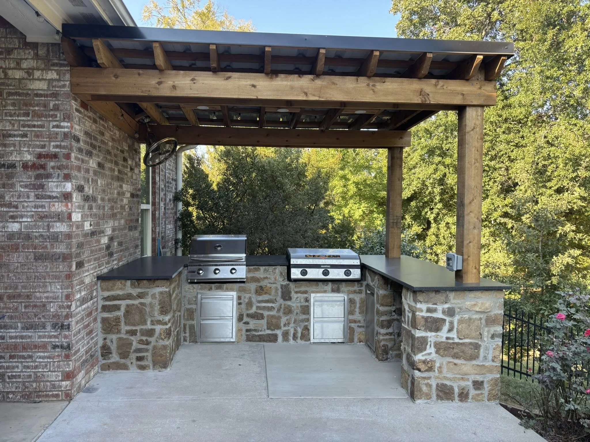 An outdoor kitchen setup with a stone base, black countertops, and a stainless steel grill under a wooden pergola with a metal roof, surrounded by greenery and trees. ETX Tyler Texas, Outdoor living, Residential New Construction