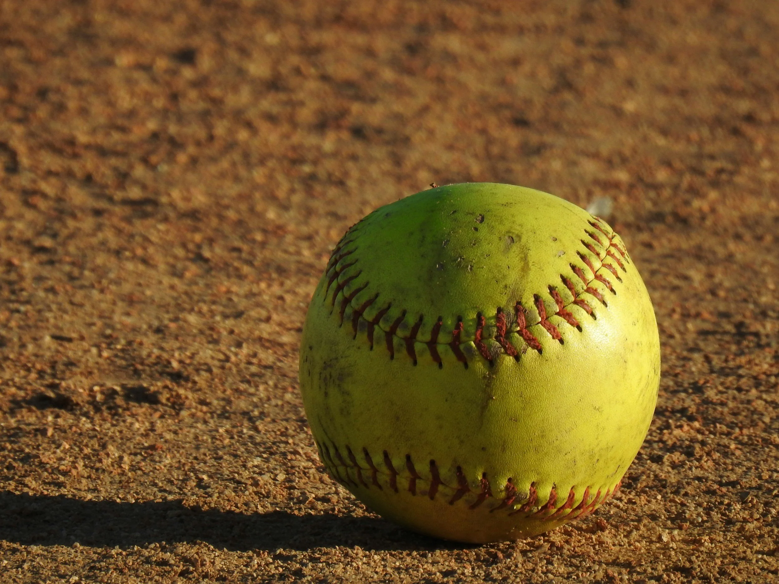 A close-up photograph of a yellow tennis ball standing upright on a sandy surface, with a shadow cast to the left and slight wear visible on the ball.