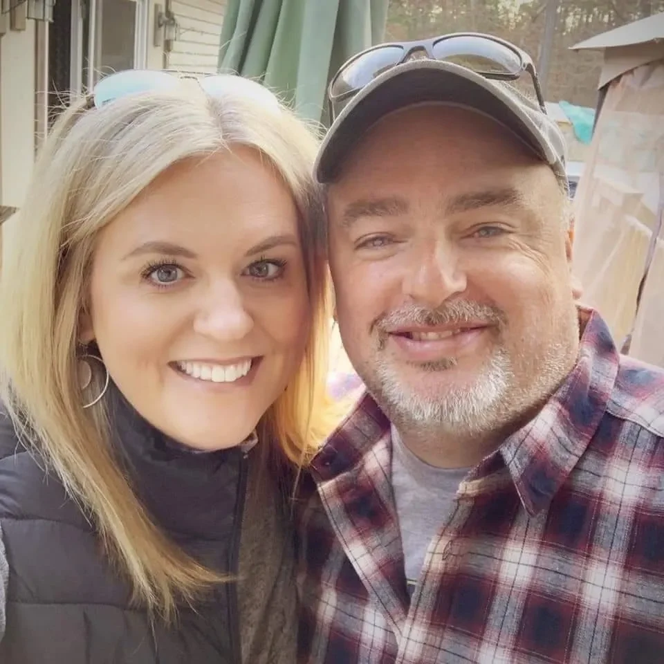 A smiling woman with blonde hair and blue eyes standing next to a man with a beard, wearing a baseball cap, who is also smiling. They are outdoors with a house and shed visible in the background.