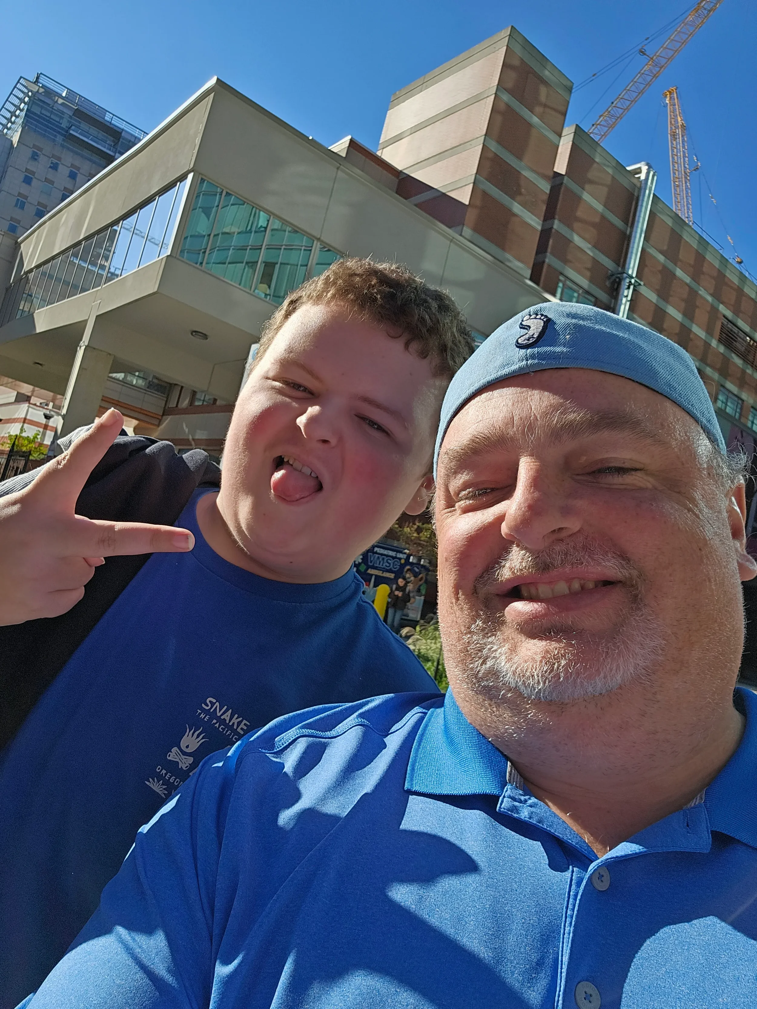 A selfie of a young boy making a peace sign and sticking out his tongue with an older man with a gray beard smiling, both outdoors in front of a modern building with glass windows and a construction crane in the background.