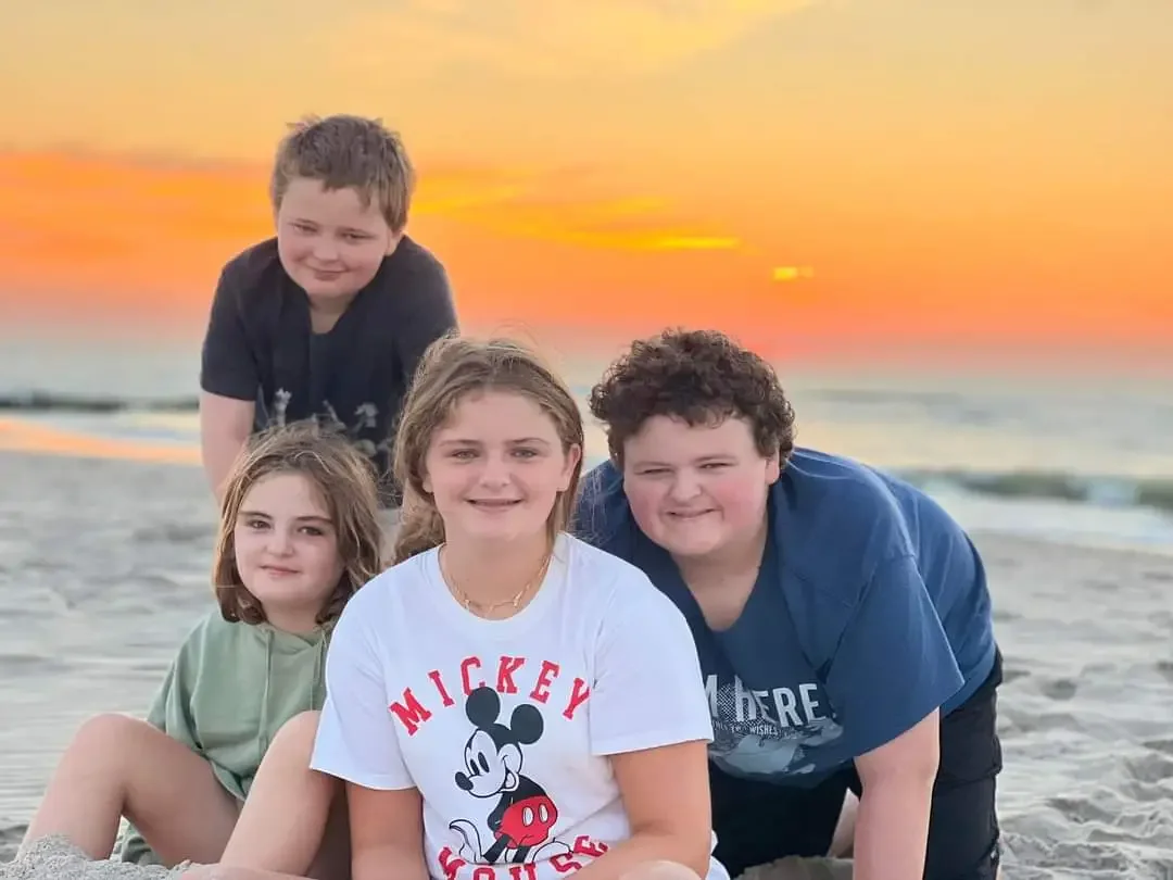 A group of five children sitting on the beach during sunset. Two boys and three girls, with one girl wearing a Mickey Mouse t-shirt.