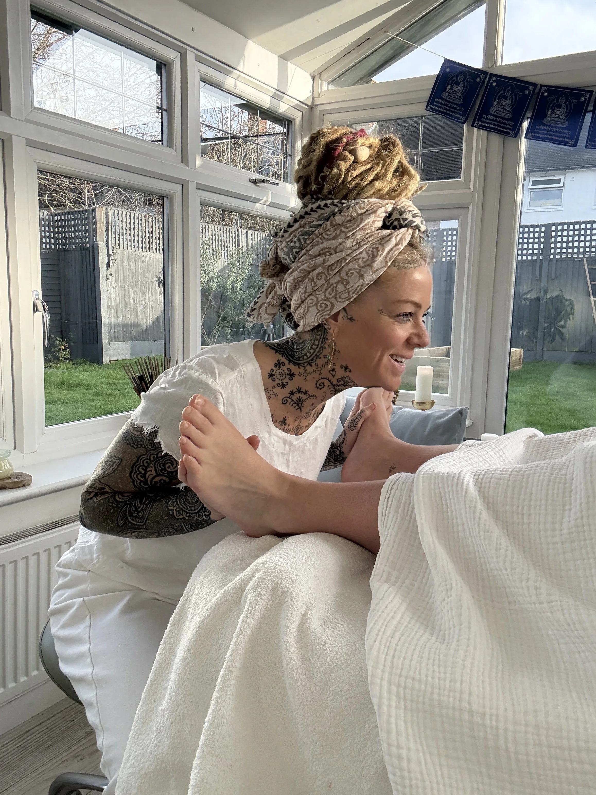 A woman with dreadlocks and tattoos is smiling and interacting with someone lying in bed, in a bright room with large windows showing a backyard.