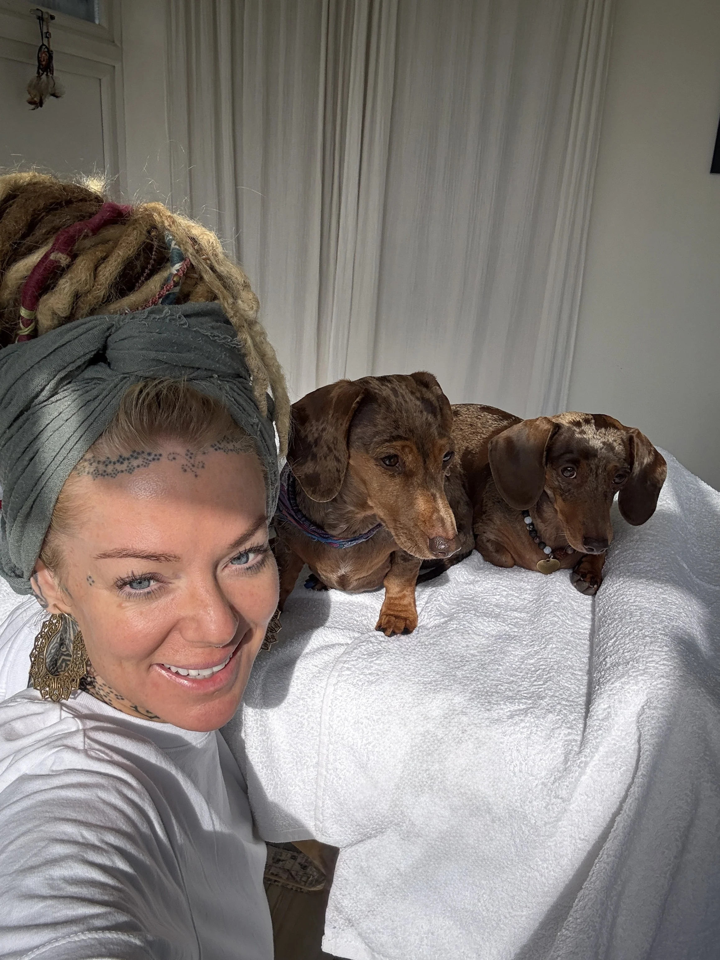 A woman with blonde dreadlocks and face tattoos smiling, taking a selfie with two Dachshund dogs lying on a white blanket on a bed.