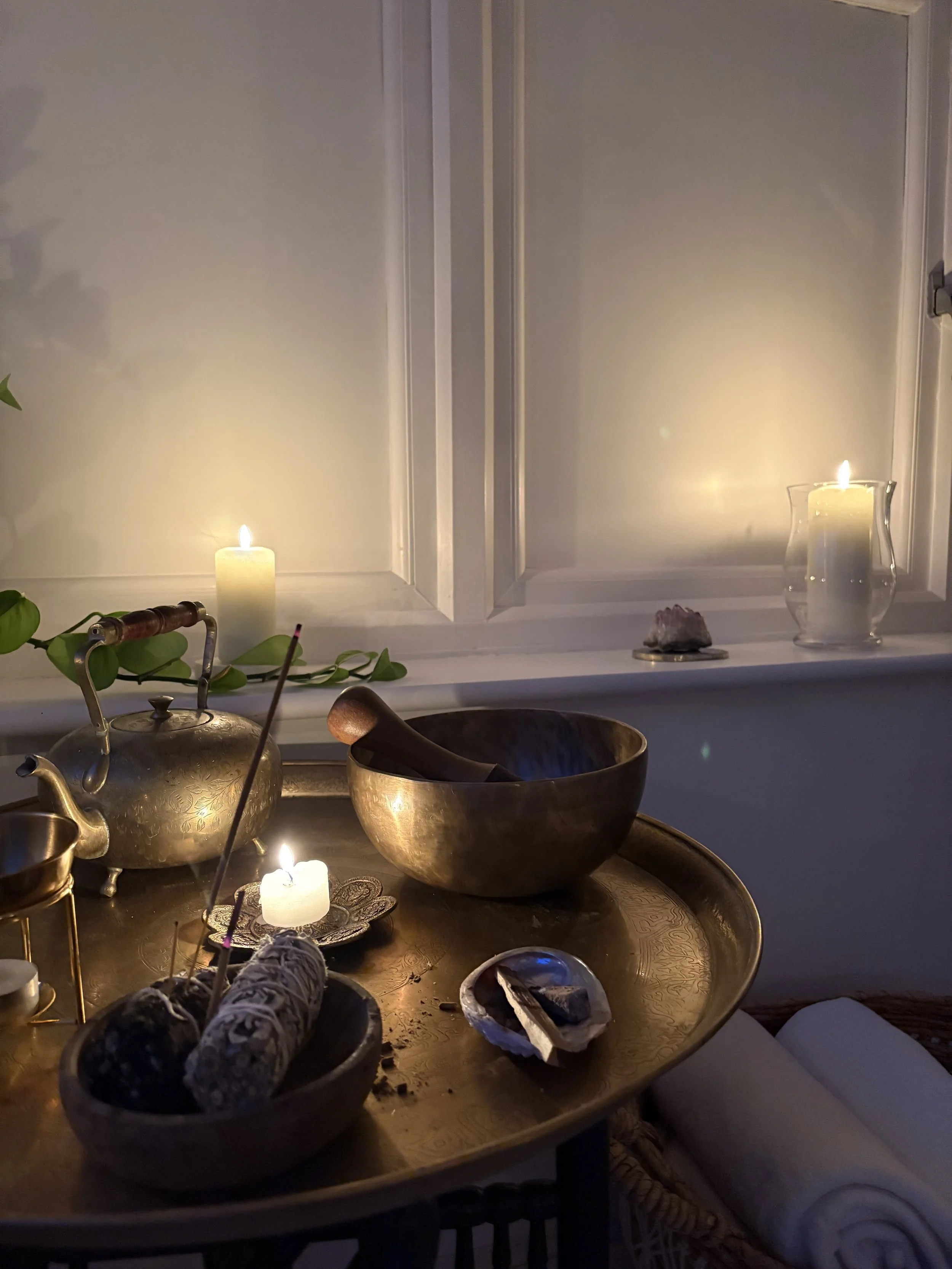 A cozy interior scene with a wooden tray on a table holding burning sage smudges, a small bowl, and other decorative items. Behind the table, lit candles and a glass pitcher are on a windowsill, creating a warm ambiance.