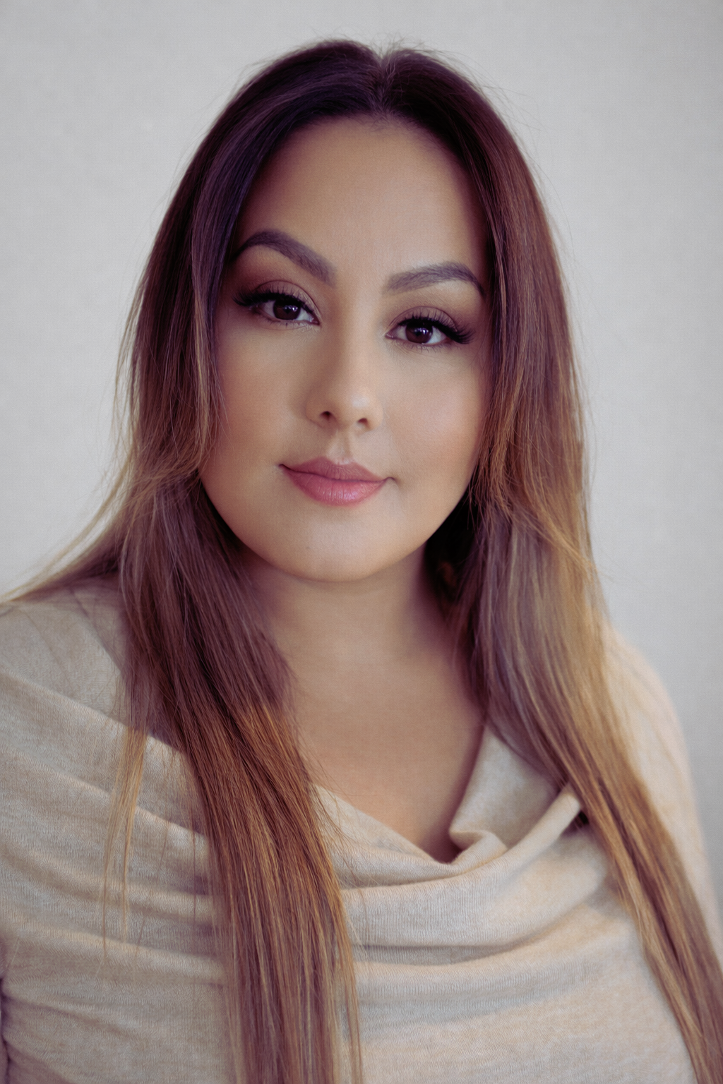 Close-up portrait of a young woman with long, wavy, auburn hair, soft makeup, and a light beige top, looking at the camera with a neutral expression.
