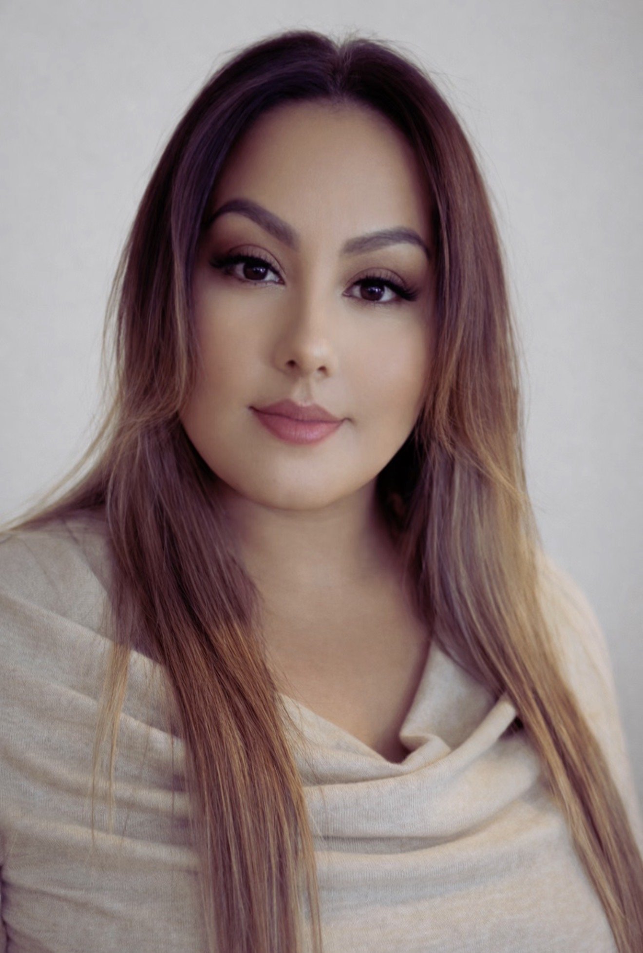 A woman with long, wavy, reddish-brown hair, wearing a beige hoodie, looking at the camera with a slight smile, against a neutral background.
