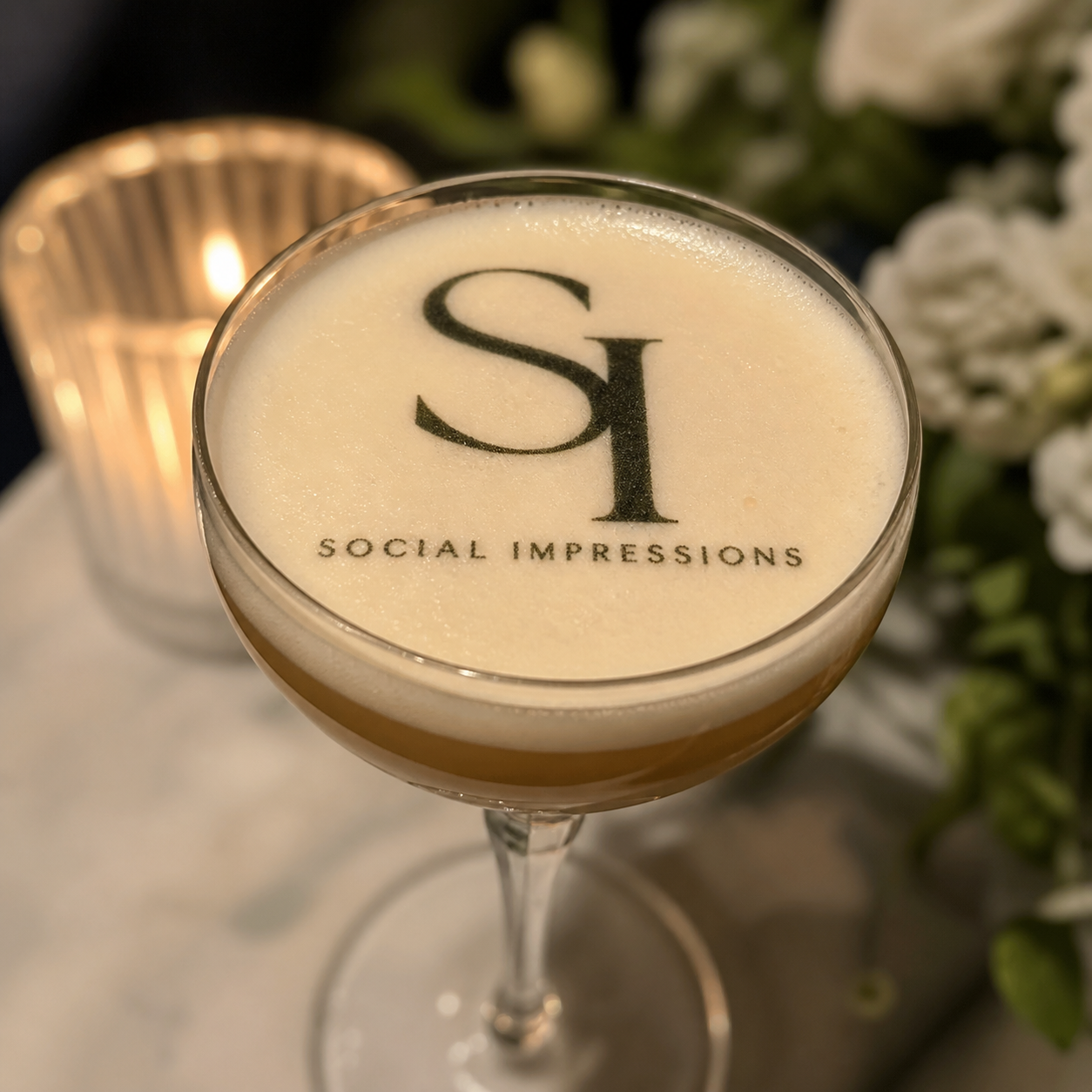A cocktail glass with a creamy drink and the logo 'SI' along with the text 'Social Impressions' on top, in a setting with a candle and flowers in the background.