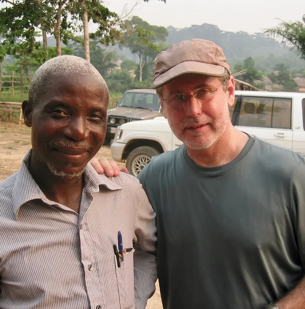 Two men stand smiling for a photo outdoors, with vehicles and trees in the background