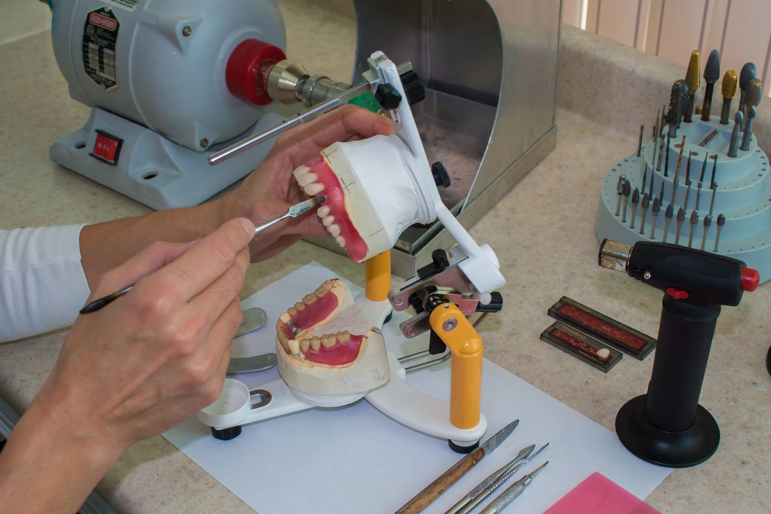 A person working on a dental prosthetic model using dental tools and equipment, with various dental instruments and a small grinding machine on a countertop.