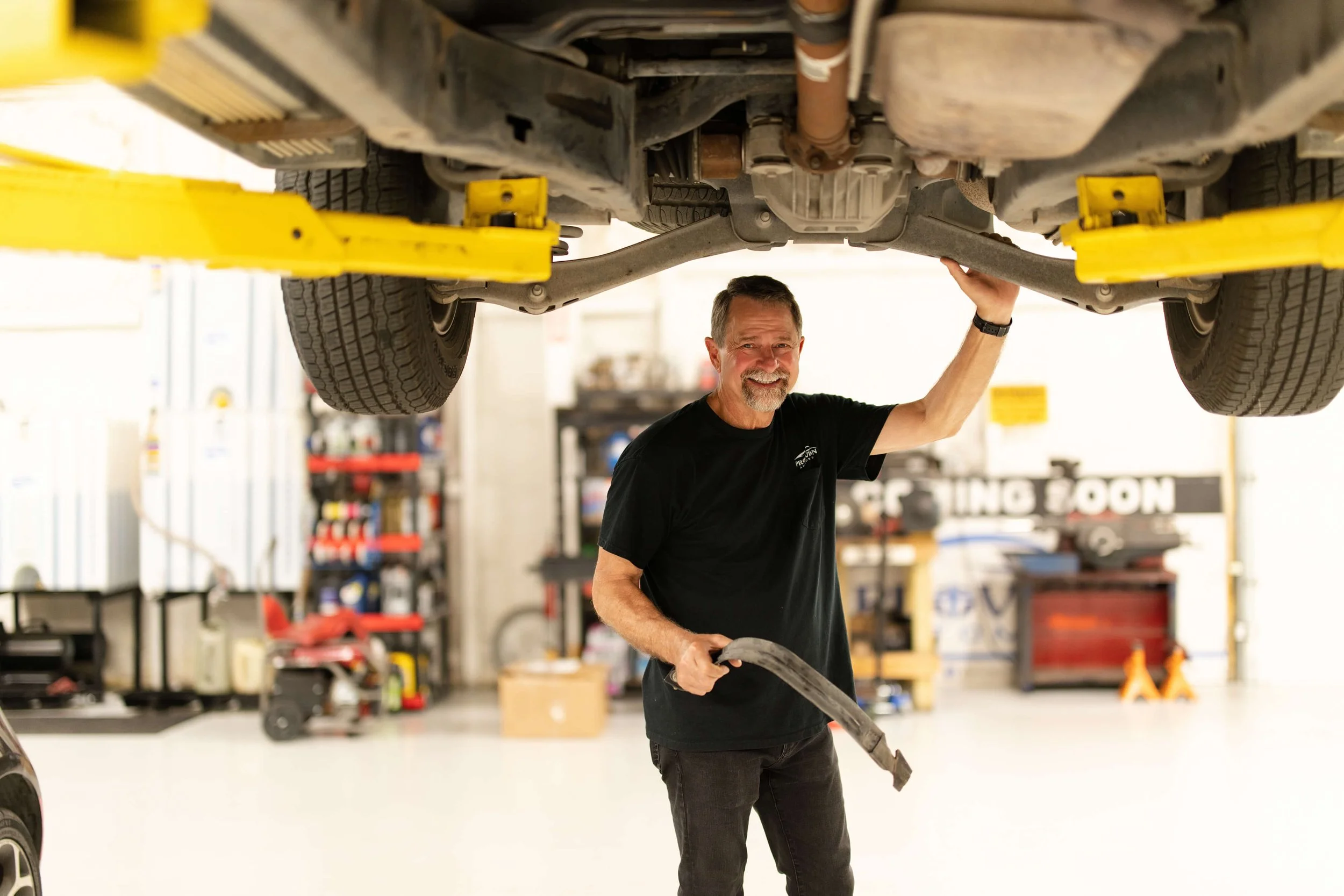 A man working underneath a vehicle that is lifted on a hydraulic lift in a garage or workshop, with tools and equipment visible in the background.