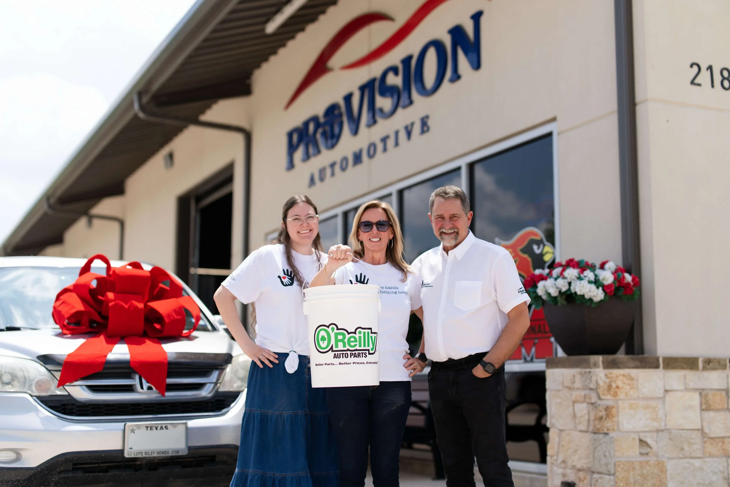 Three people stand in front of a car at an automotive store, with a large red bow on the hood, holding a donation bucket with 'O'Reilly Auto Parts' logo, in front of aProvission Automotive business, with flowers and signage visible.