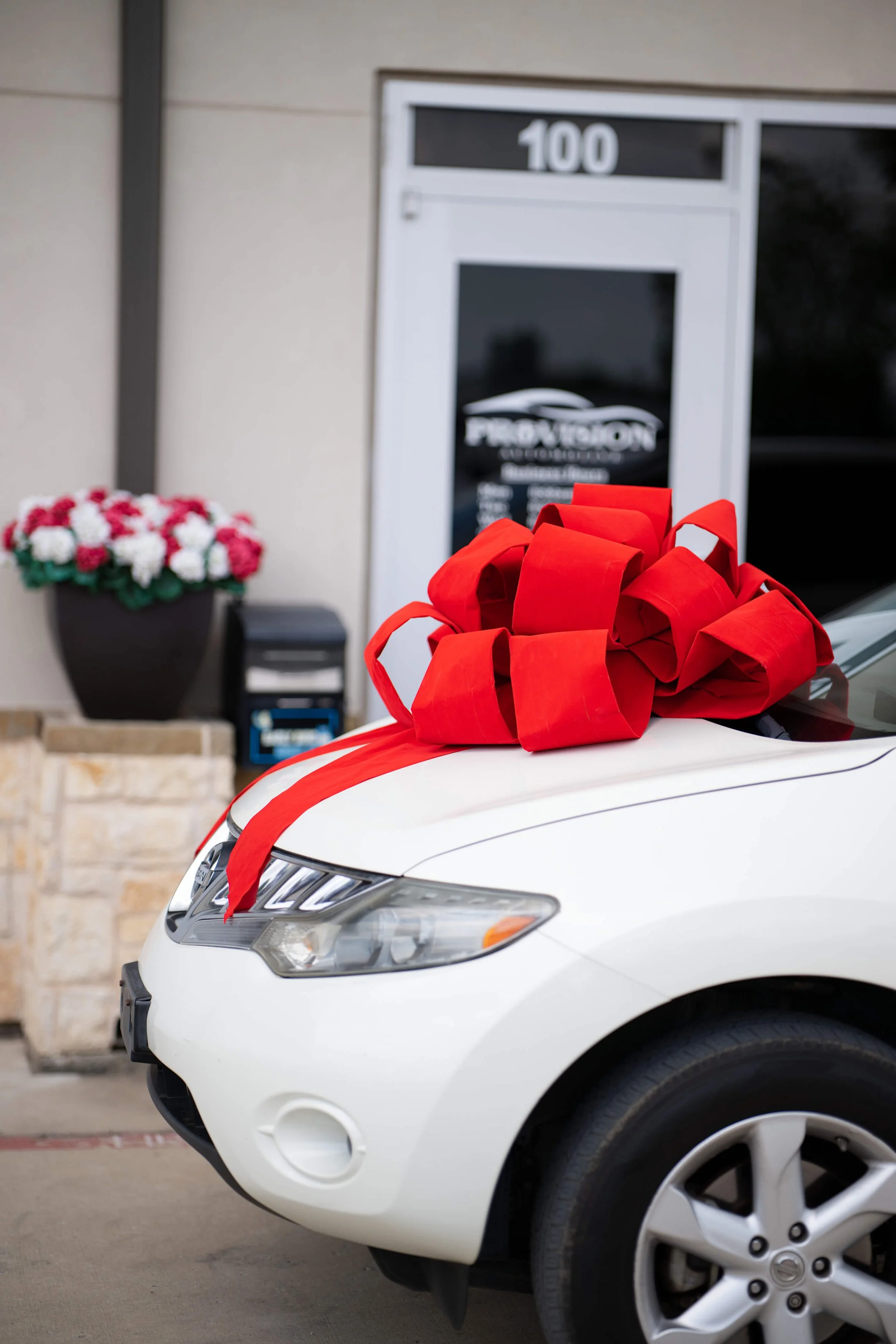 White car decorated with a large red ribbon, parked in front of a building with a flower pot containing pink and white flowers, and a sign with the number 100.