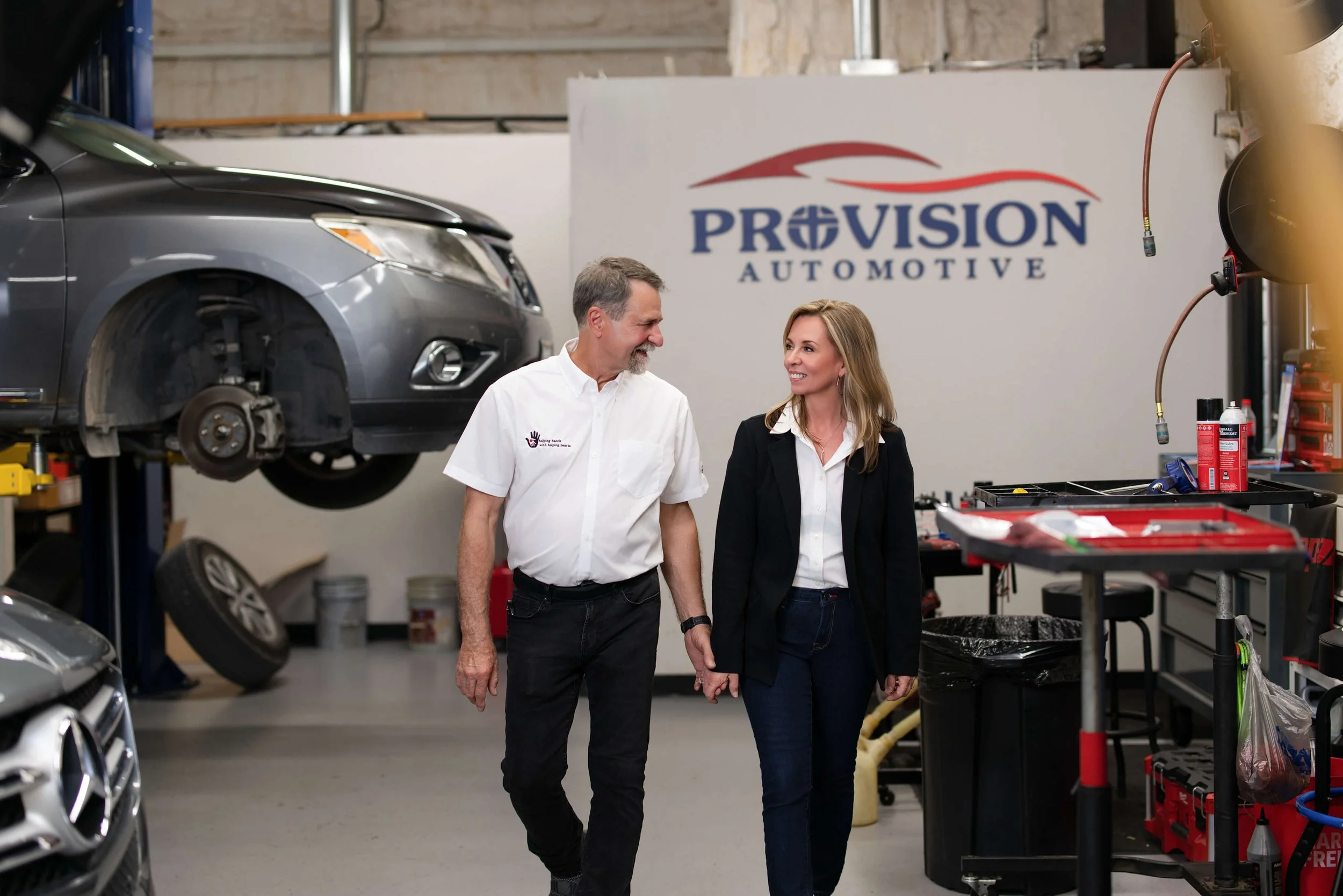 Two people, a man and a woman, are walking through an automotive repair shop, smiling and holding hands. The shop has a car on a lift with its front wheel removed, and various tools and equipment are visible.