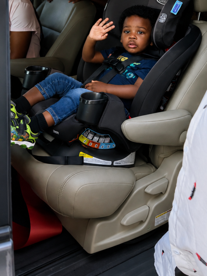 A young boy in a car seat inside a vehicle, waving. An adult man stands nearby, partially visible. Other children are in the background, some smiling and peeking through the headrest of the seat.
