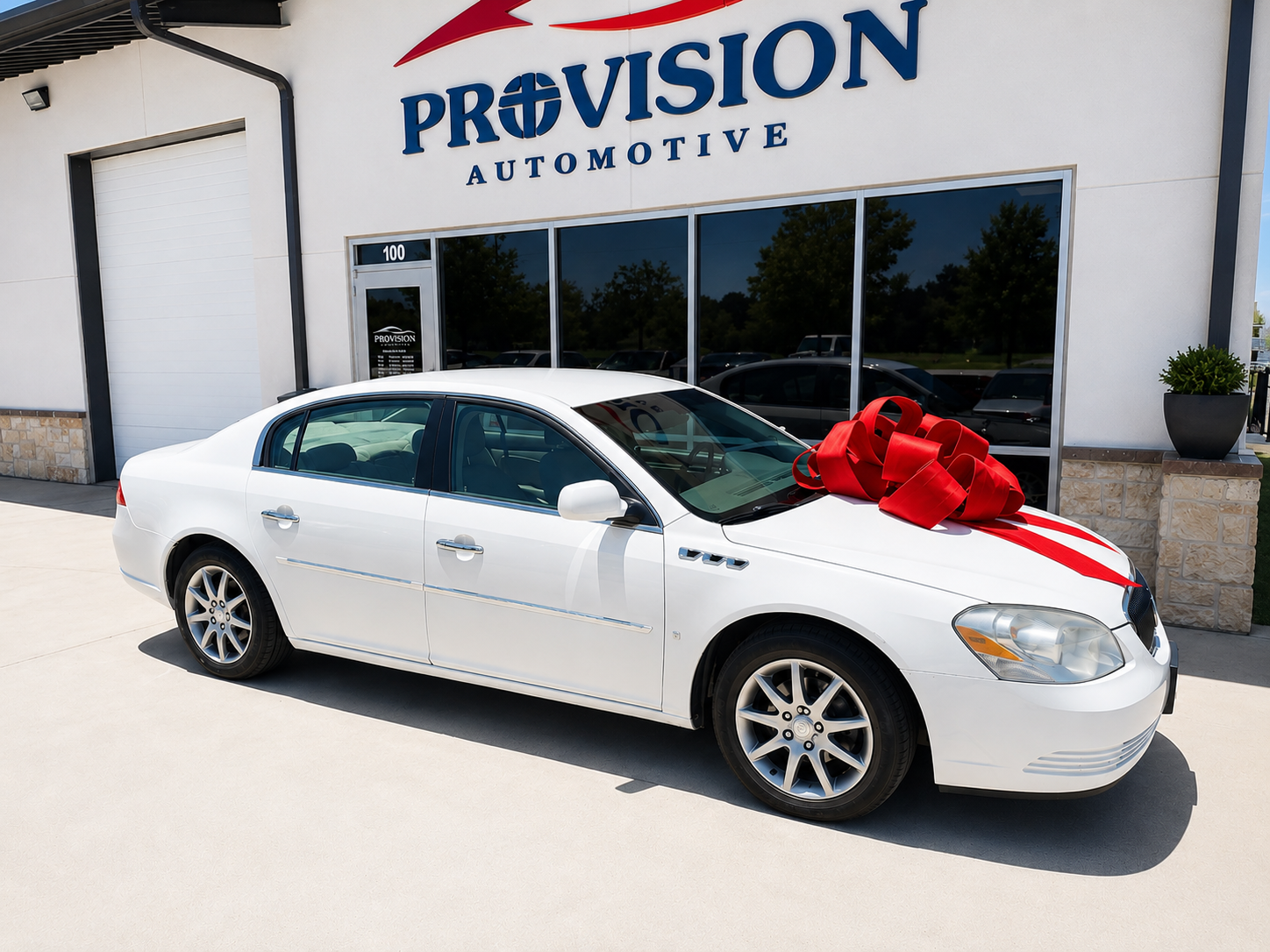 White sedan car with a large red ribbon and bow on the hood, parked outside Provision Automotive dealership.