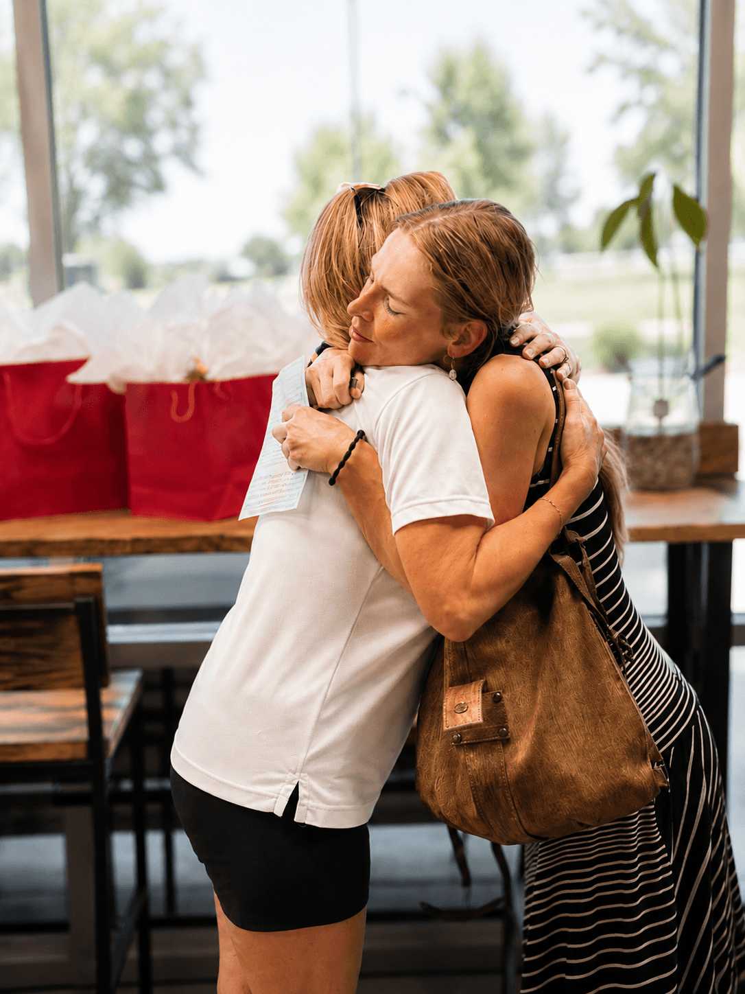 Two women hugging each other warmly in a bright room with windows in the background. One woman with short hair is wearing a white shirt and black shorts; the other has long hair and is wearing a black and white striped dress and carrying a brown bag.