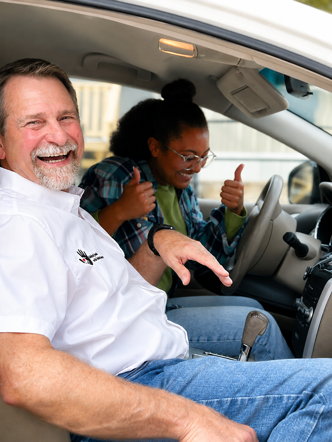 Two people sitting in a car, both smiling and showing thumbs up. The man in the front seat has a white beard and is wearing a white shirt. The woman in the back seat has glasses, curly hair, and is wearing a plaid shirt.