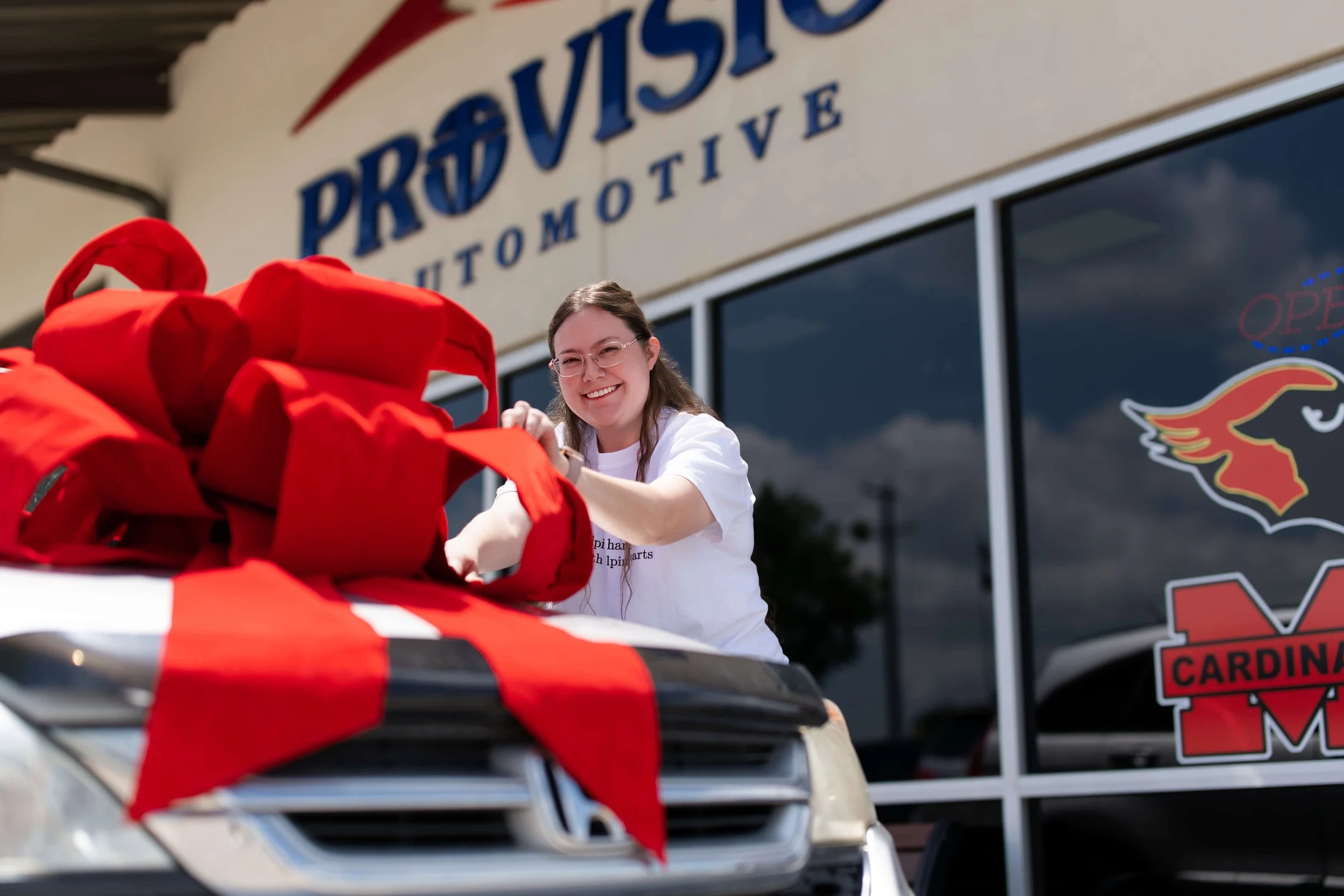 A woman with glasses and a white shirt smiling as she prepares a red gift bow on a vehicle's hood, in front of an automotive store sign that reads 'PROVO AUTO'. The scene is outdoors with the store's window and door visible behind her.