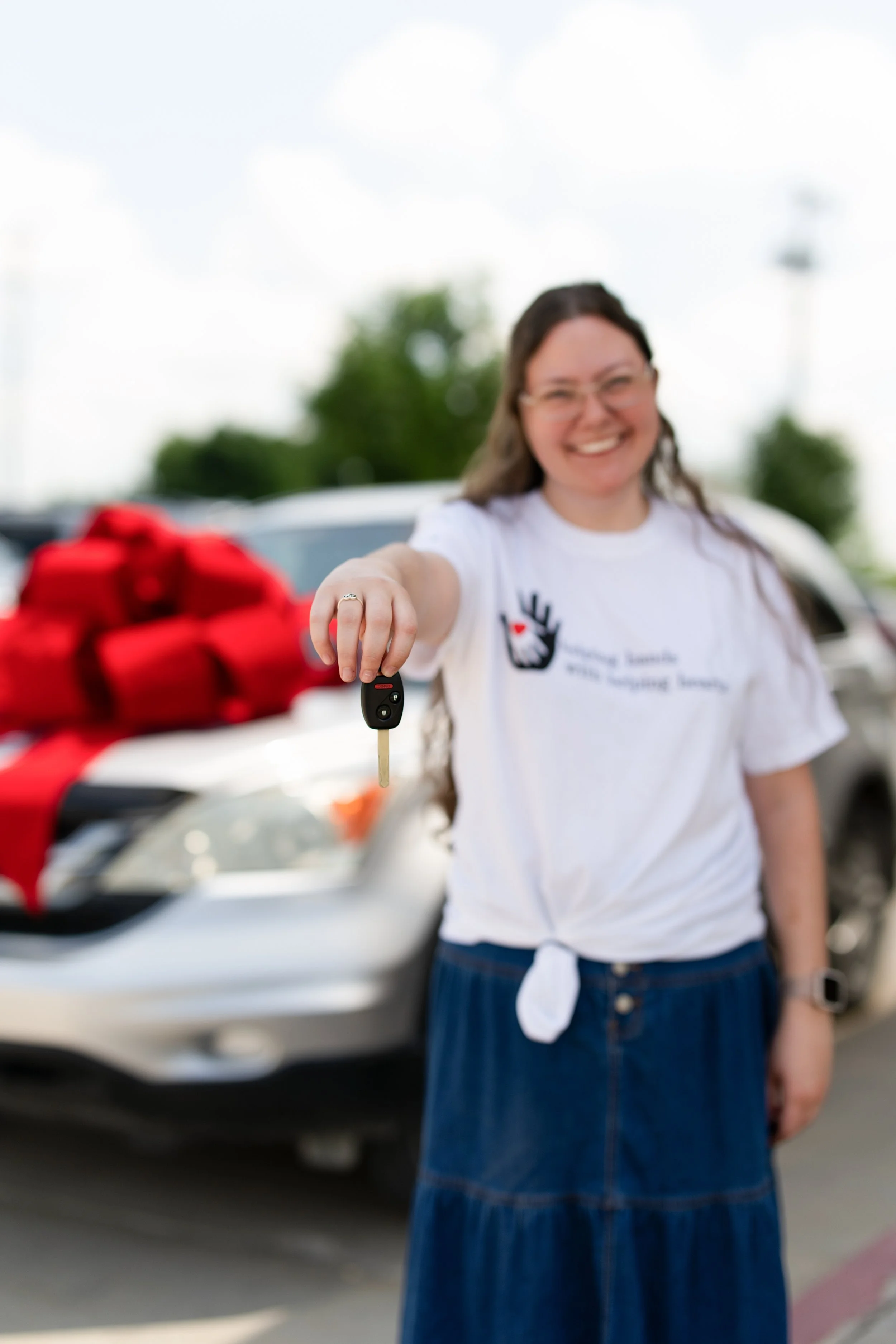 A woman smiling and extending her hand, holding a car key, in front of a white car with a red ribbon on it, outdoors in a parking lot with trees in the background.