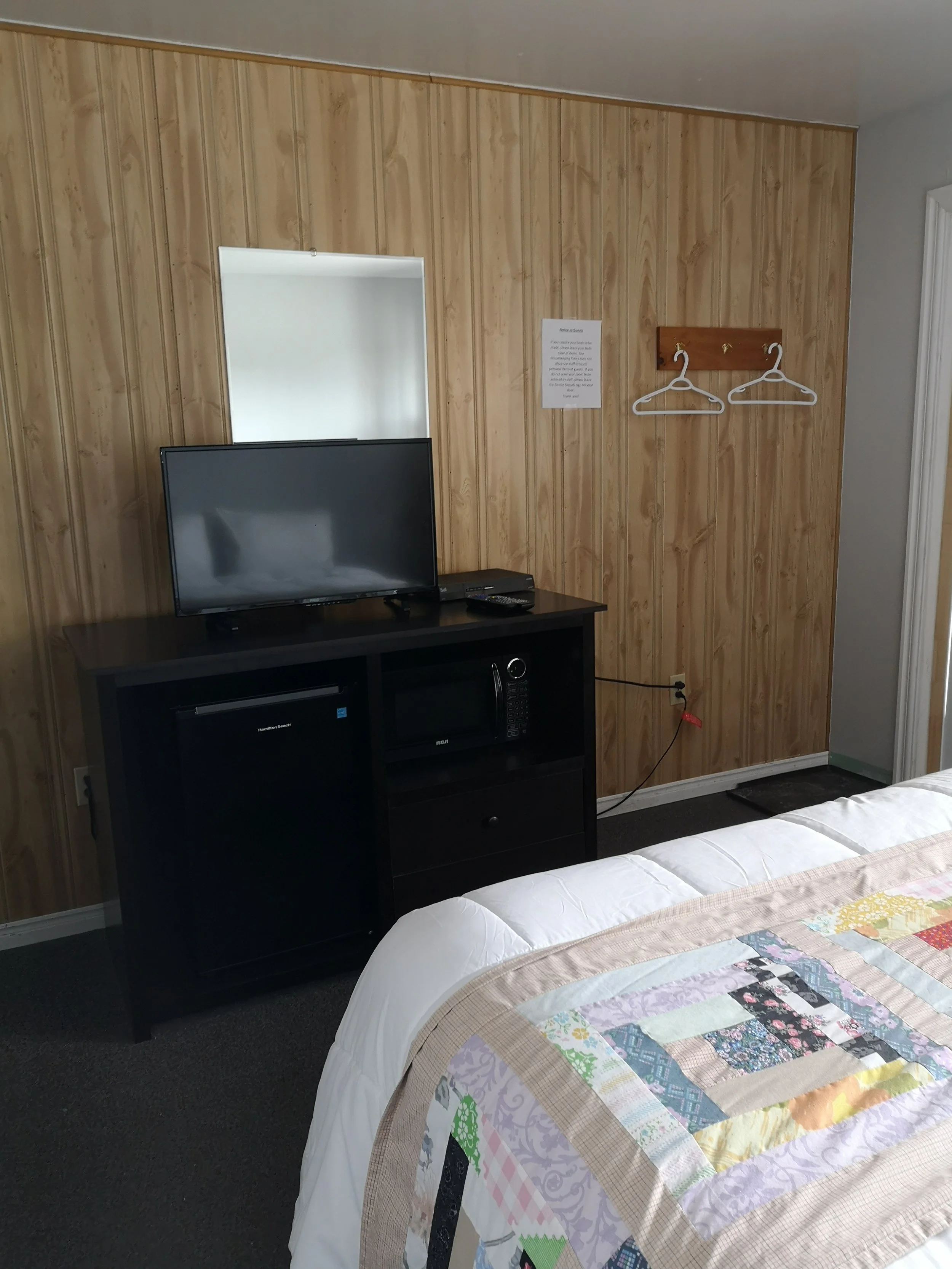 A bedroom with wood-paneled wall, a black TV on a stand, and a quilted bedspread with various pastel-colored patterns.