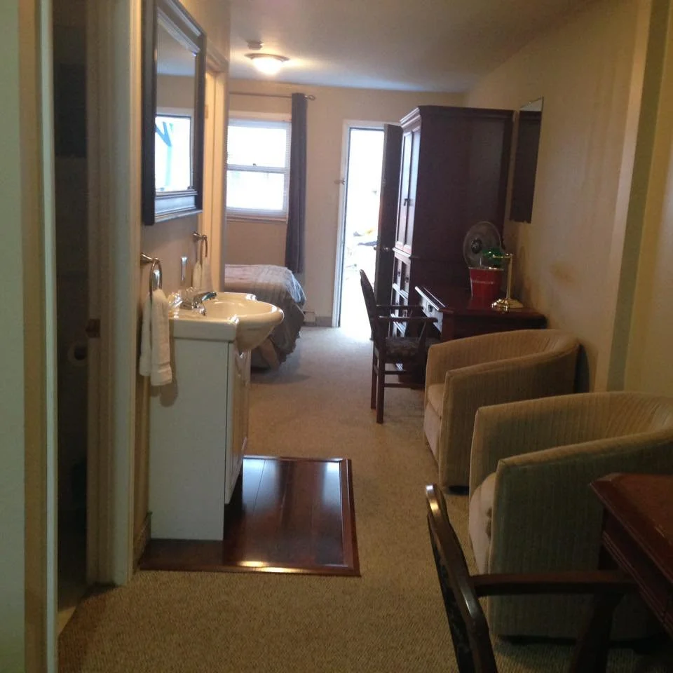 A cozy room with a small bed near the windows, a white sink with a towel, a wooden cabinet, and a beige armchair, illuminated by natural light from the windows and a ceiling light.