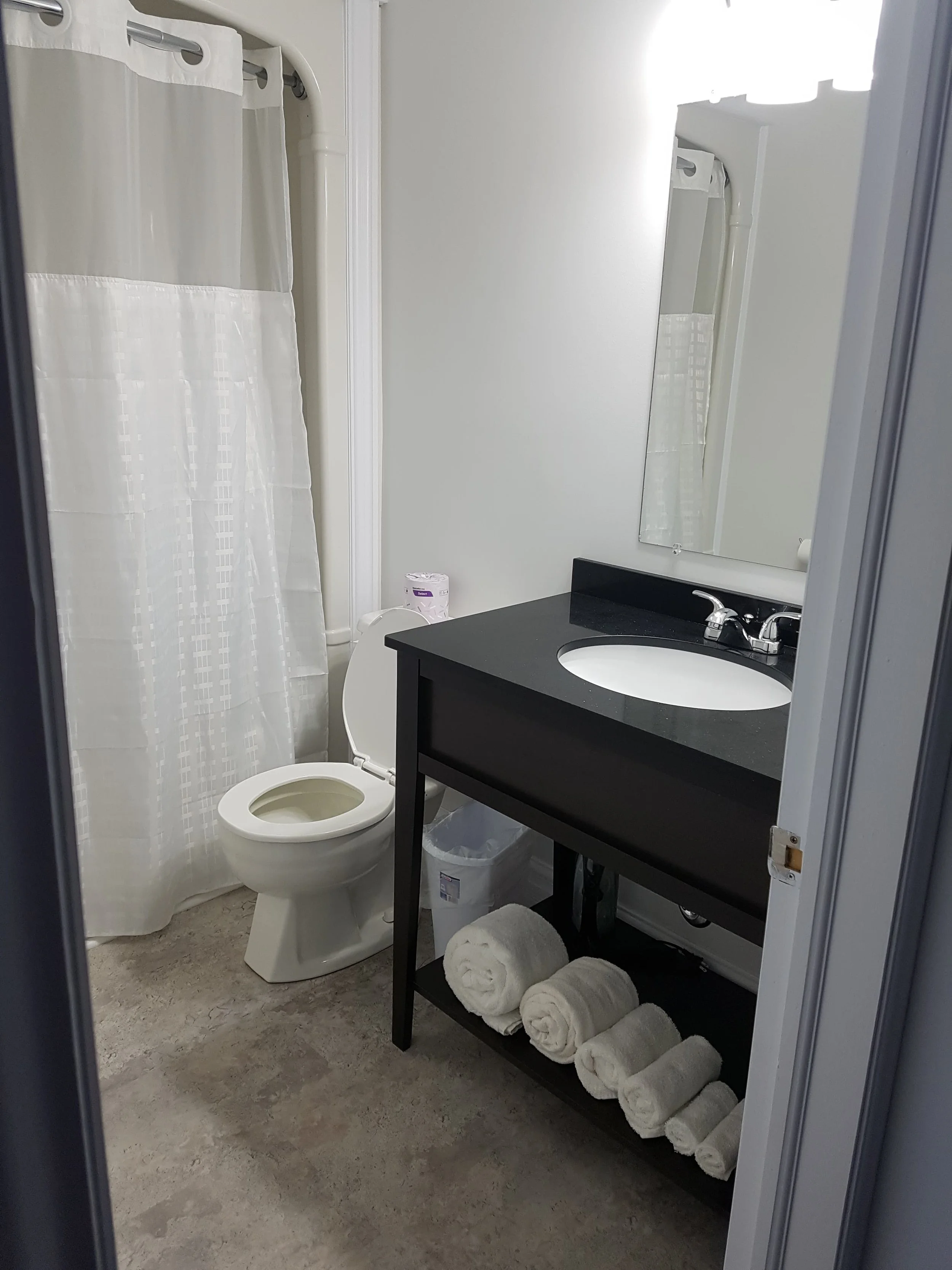 A small bathroom with a white toilet, a black vanity with a sink, a mirror, white rolled towels on a shelf, and a shower with a curtain.