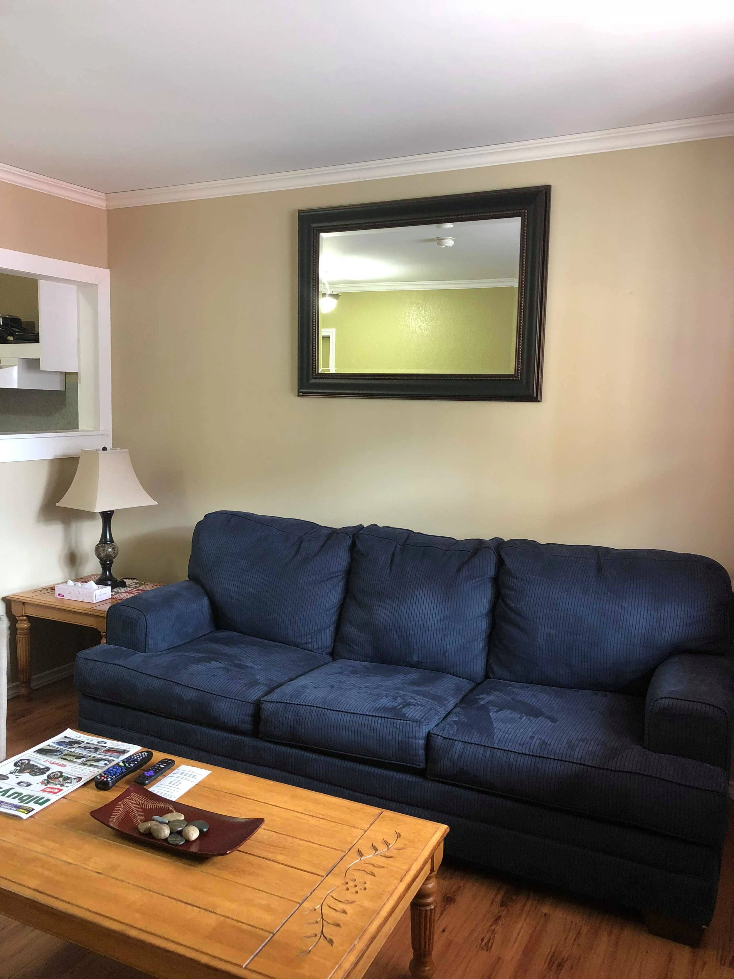 Living room with a blue sofa, wooden coffee table with decorative stones and magazines, beige walls, a large mirror on the wall, a lamp on a side table, and partial view of a kitchen counter.