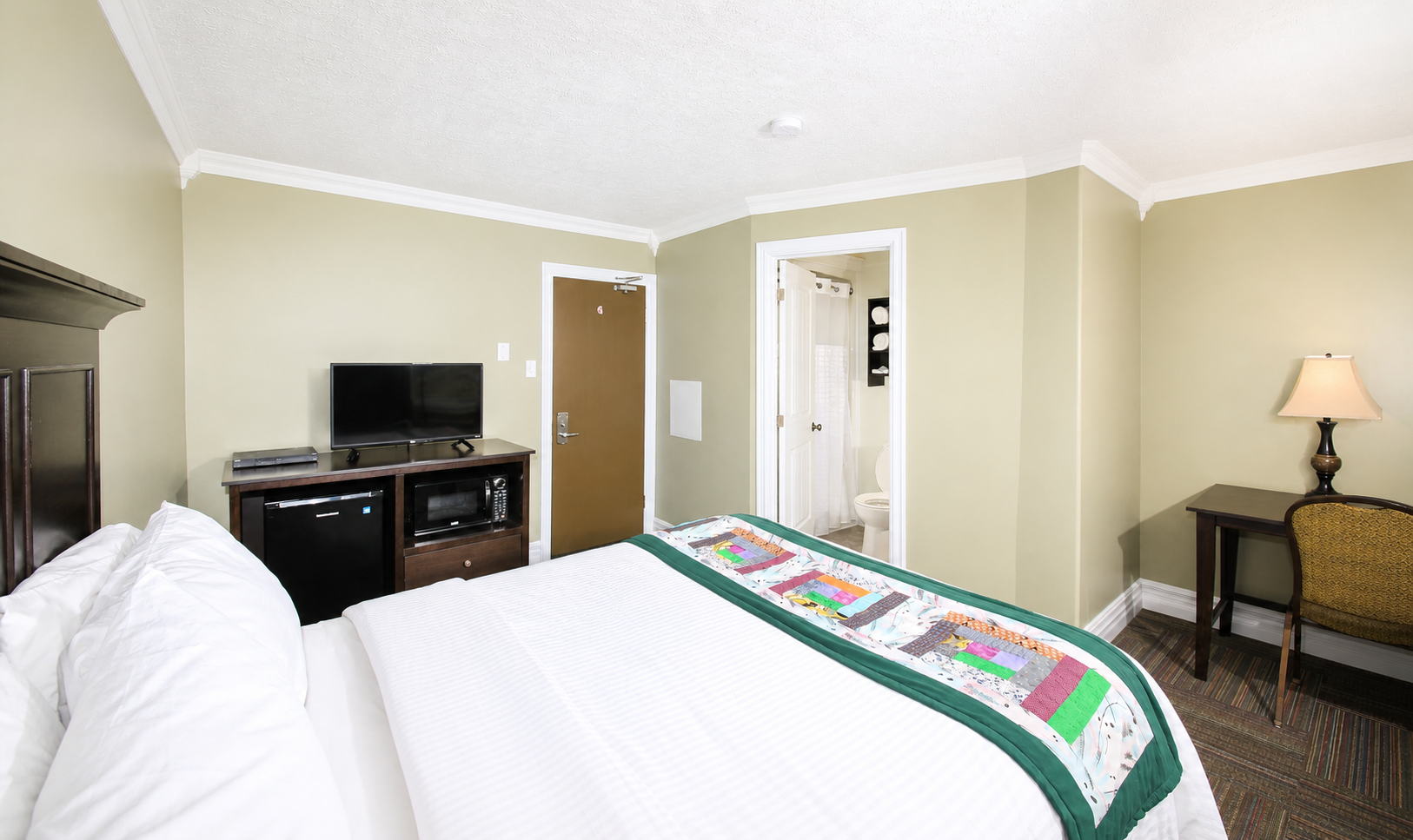 Hotel room with a bed, television on a stand, a doorway to a bathroom, a small desk with a lamp, and a window with curtains.