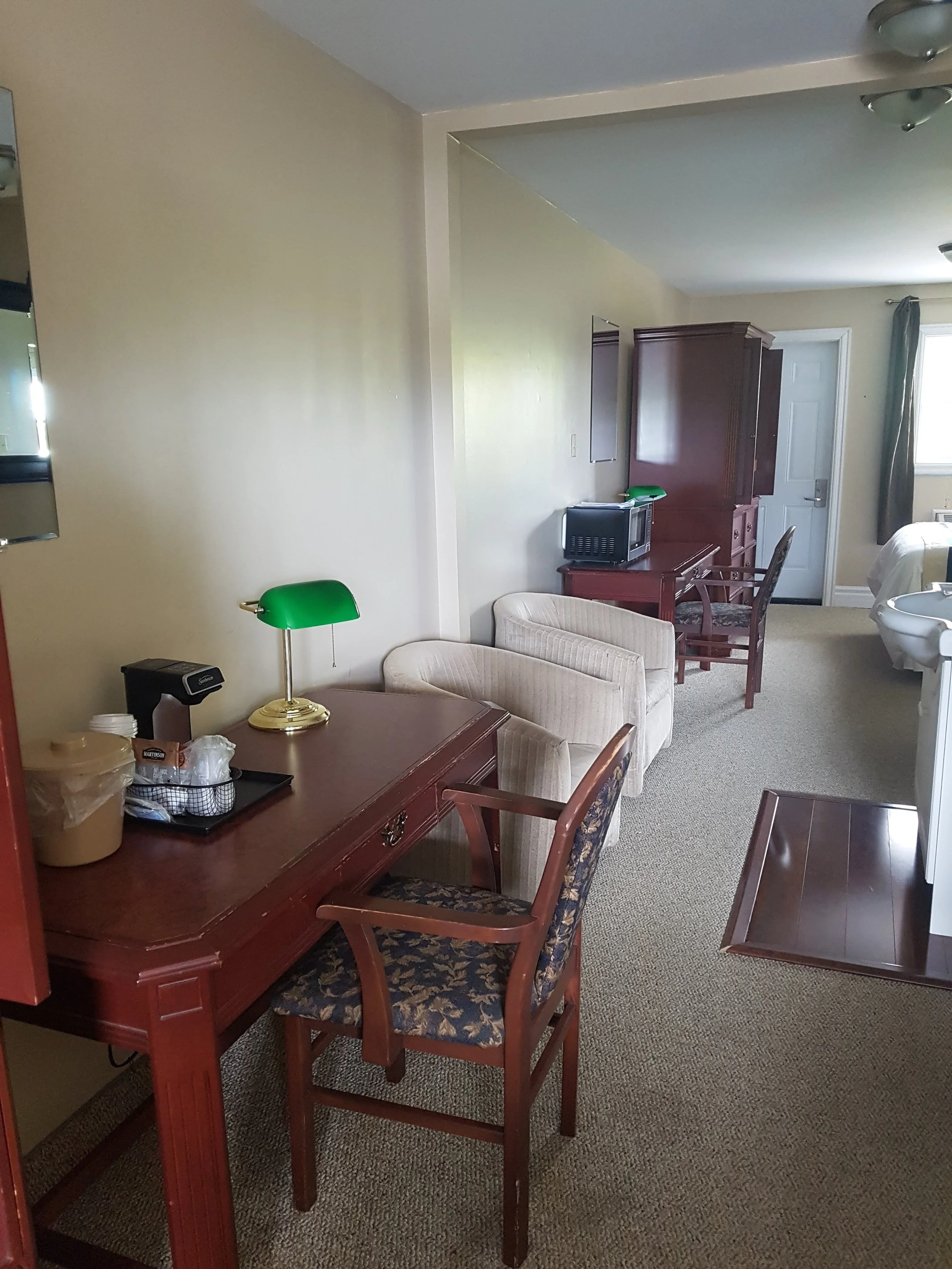 Inside a hotel room with a beige wall, a wooden dining table with floral upholstered chairs, a beige armchair, a green desk lamp, a small microwave, a TV, a white door, and a window with curtains.