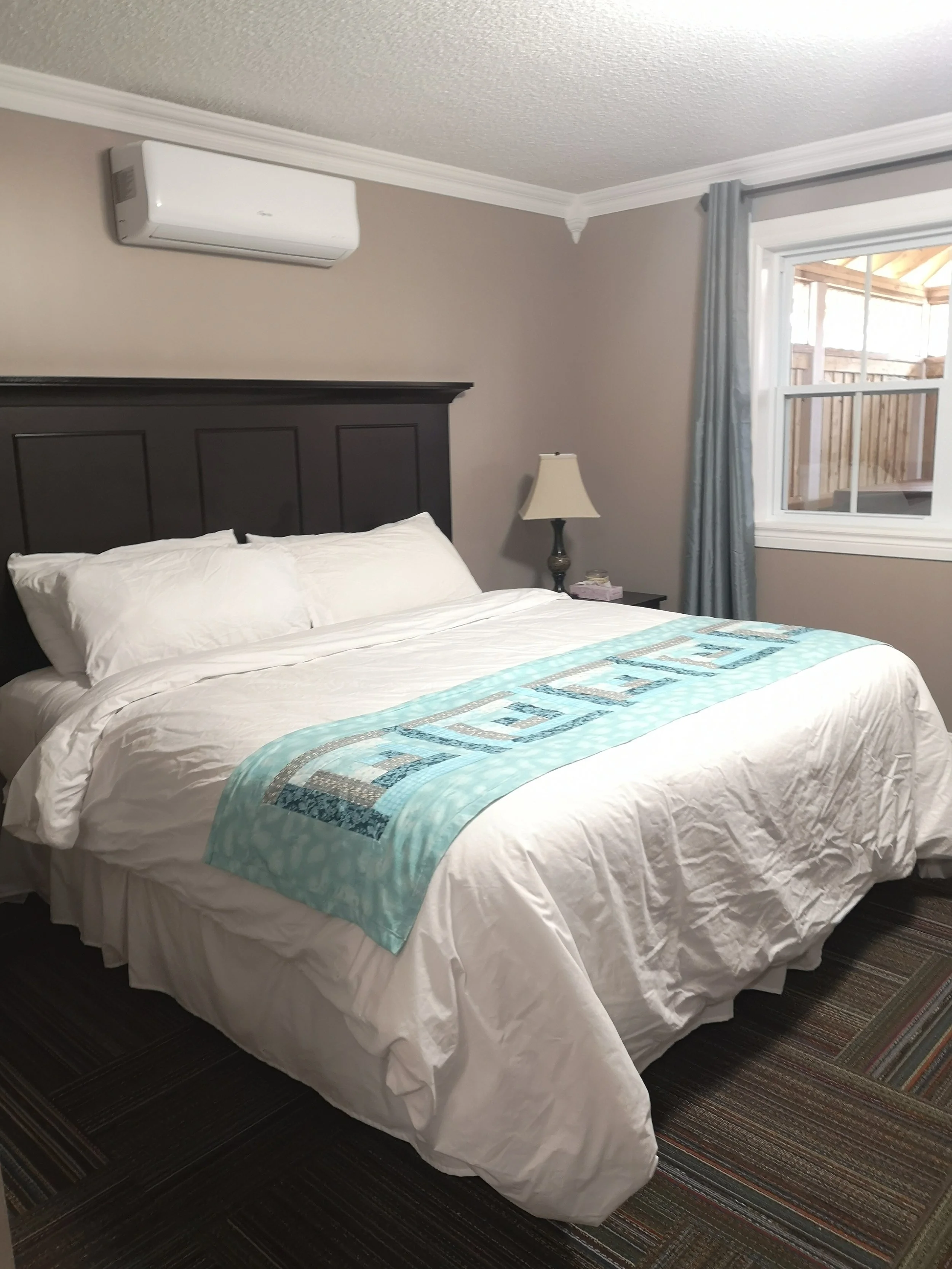 A neatly made bed with white sheets and pillows, a light blue and gray quilt, a dark headboard, a bedside table with a lamp, and a window with gray curtains.