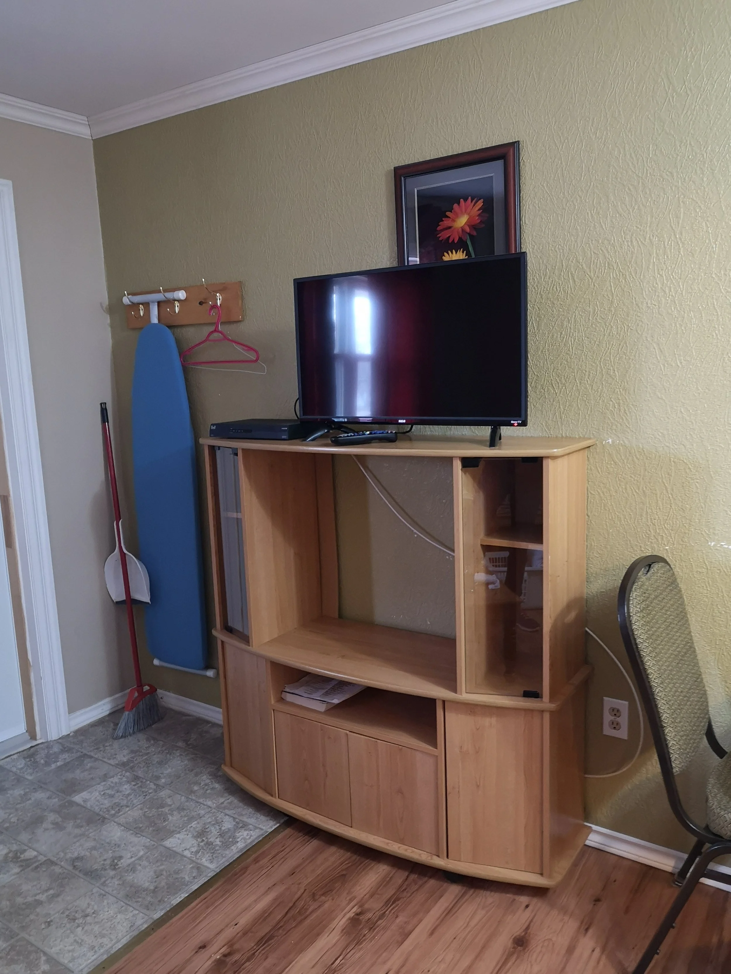 Empty wooden TV stand with a flat-screen TV on top, framed flower picture hanging above, blue ironing board and mop leaning against a green wall, part of a beige wall and a black chair with a woven seat in a room with tiled and wooden flooring.