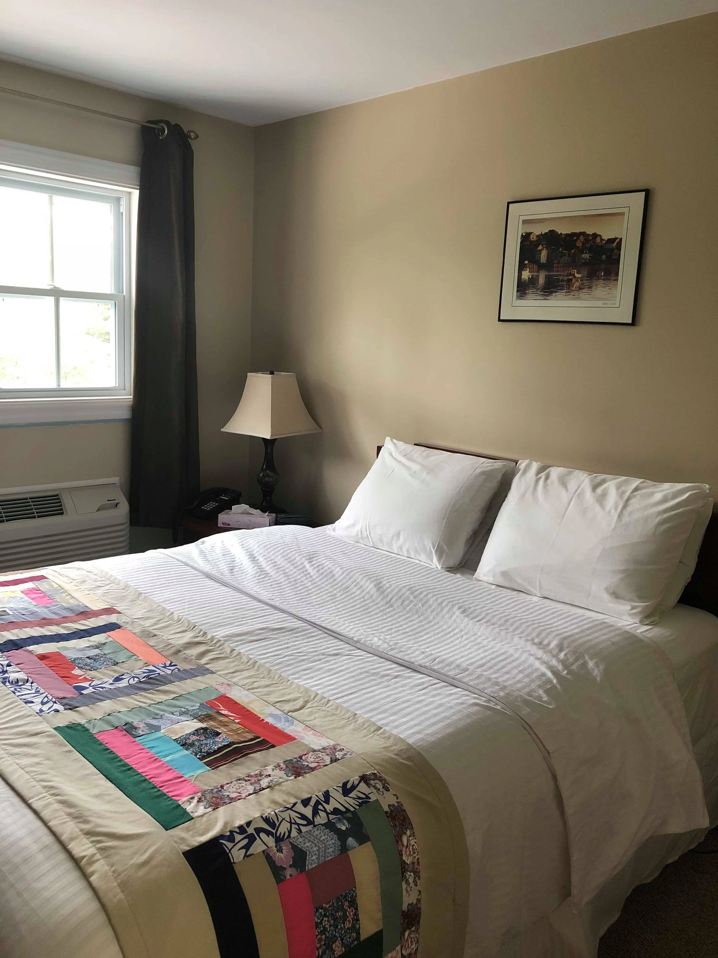 A neatly made bed with white pillowcases and a quilt featuring multicolored patches, next to a window with dark curtain. A framed picture of a seaside town hangs on the beige wall. A bedside table holds a lamp, a telephone, and some papers.