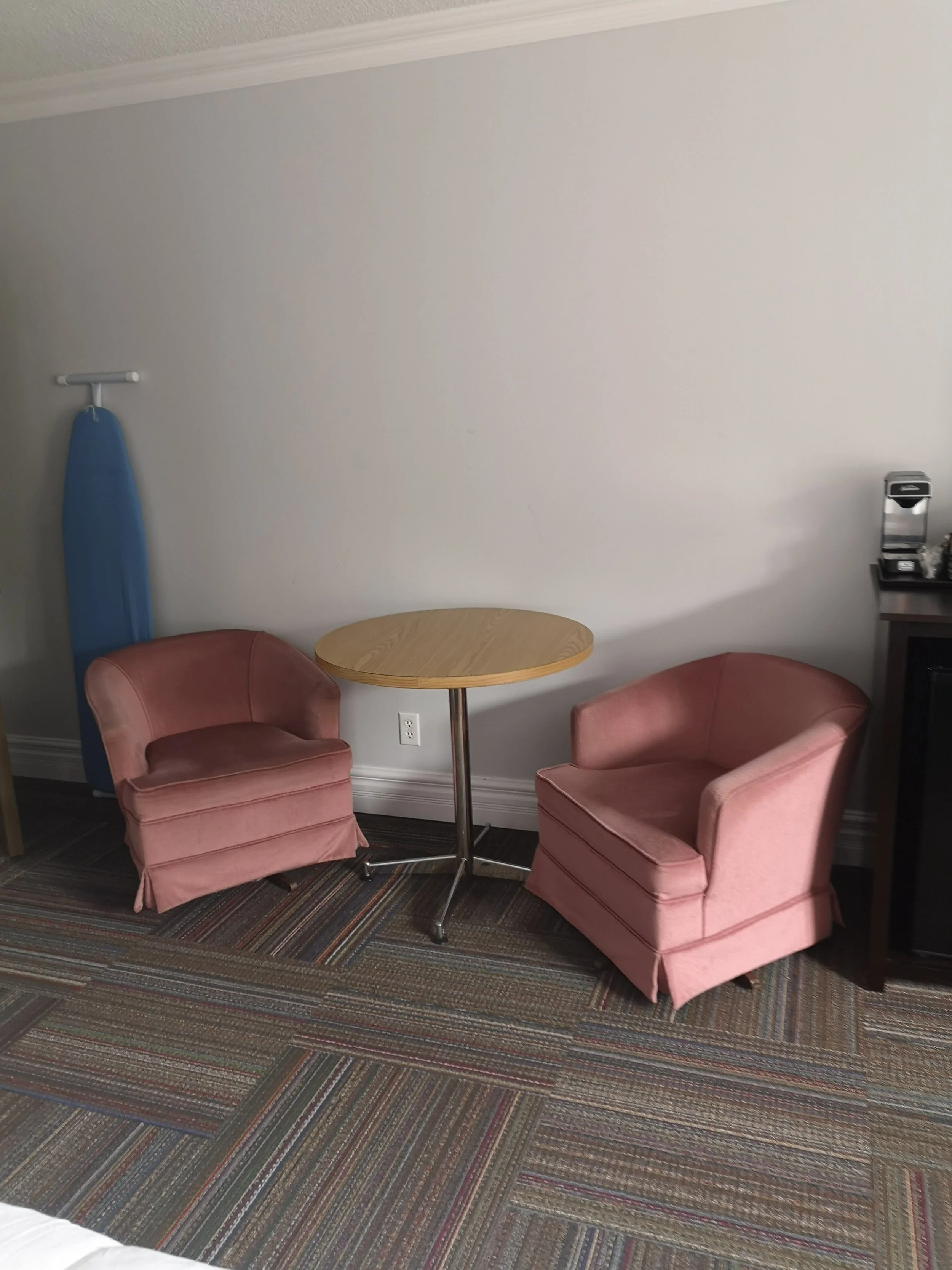 A hotel room corner with two pink armchairs around a small round wooden table, an ironing board with a blue cover against the wall, a patterned carpet, and a small countertop with a coffee maker and other items on it.