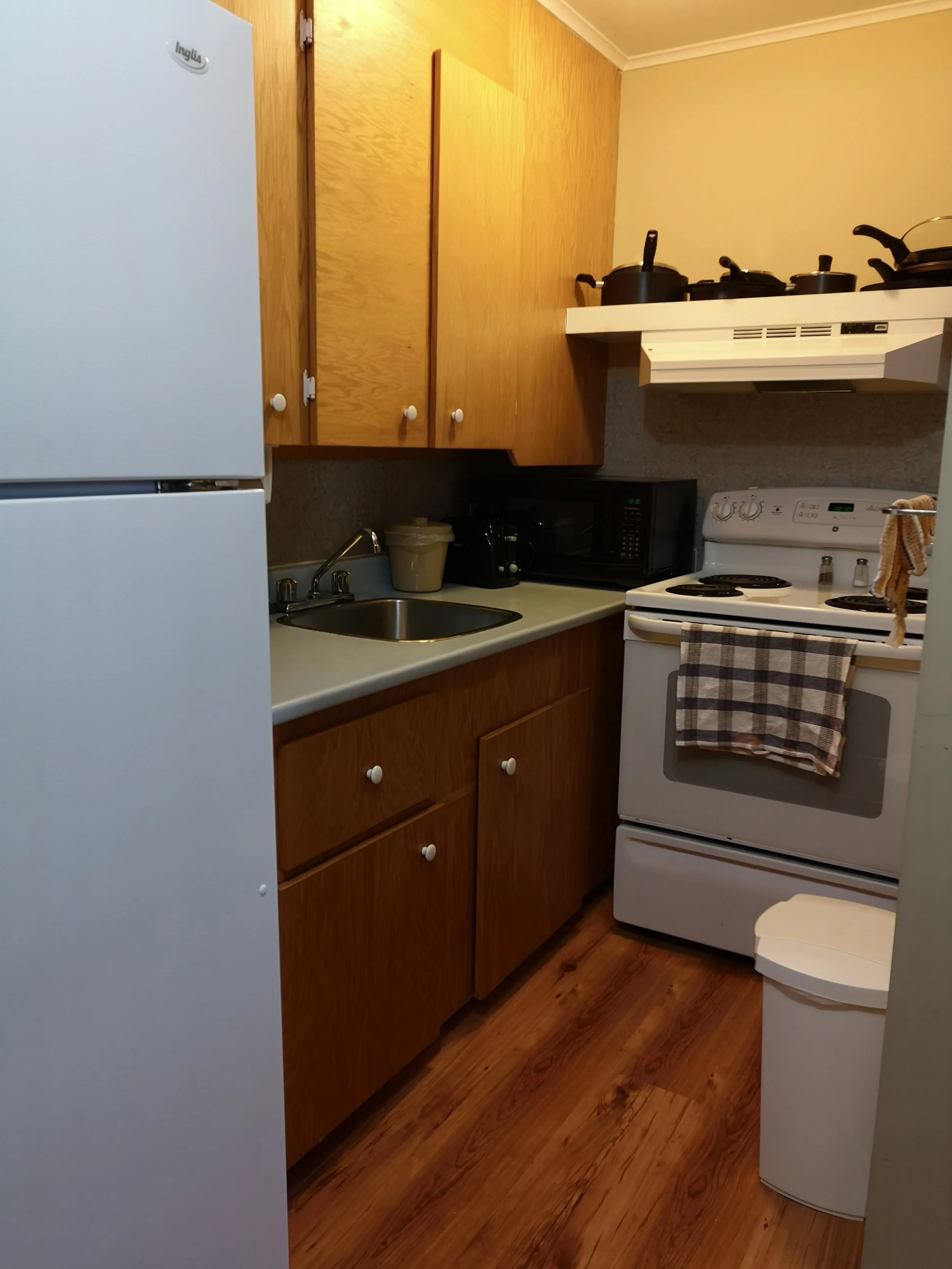A small kitchen with wooden cabinets, white refrigerator, black microwave, white stove with a towel hanging on the oven handle, and a small trash can, with a yellow wall and wood flooring.