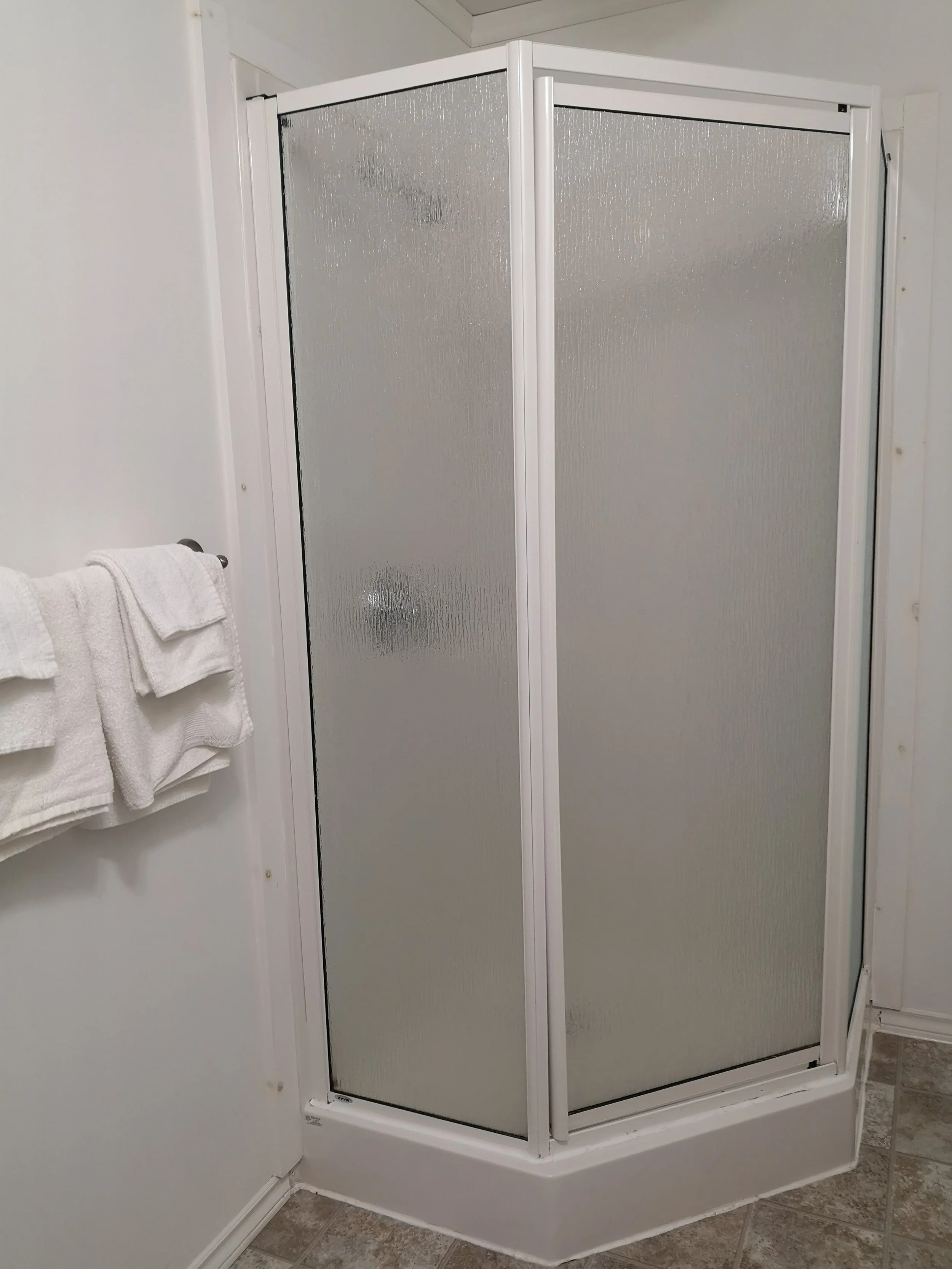 A corner shower with frosted glass panels and a white frame in a bathroom with white walls and tile flooring, with towels hanging on a rack beside it.