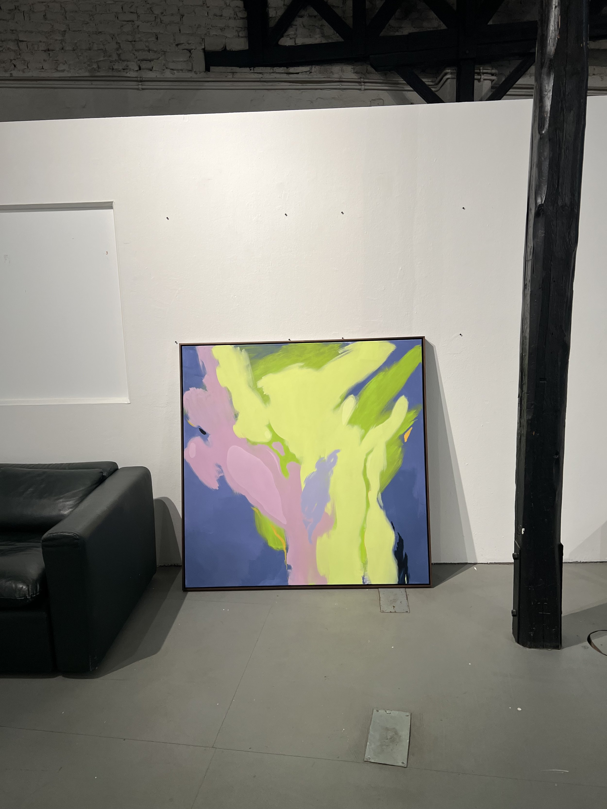 An abstract painting with vibrant colors leaning against a white wall, next to a black leather couch in an industrial-style room with exposed brick and beams.