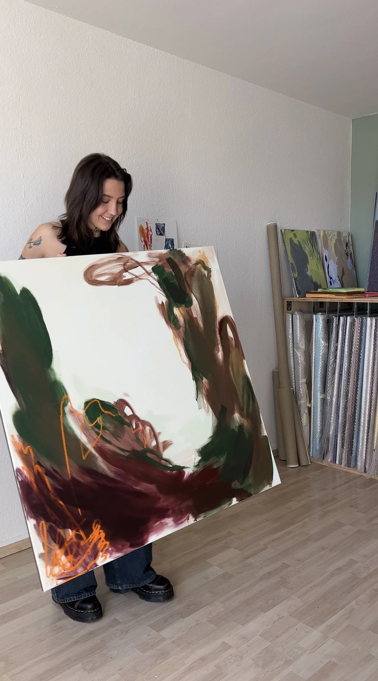 Person holding an abstract painting in a room with some other artworks and rolled canvases stored on shelves.