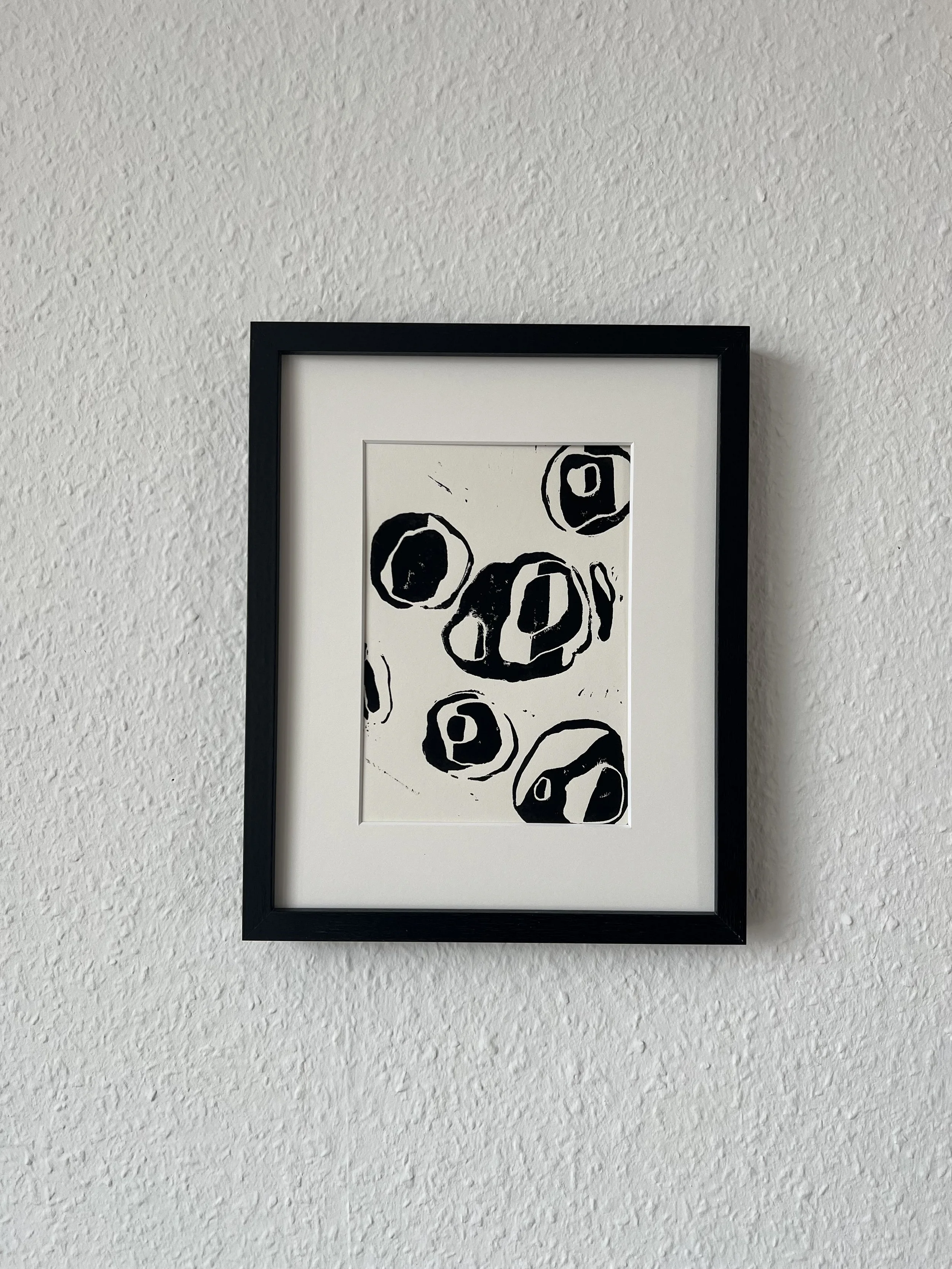 Black and white abstract artwork of circular shapes in a black frame on textured white wall.