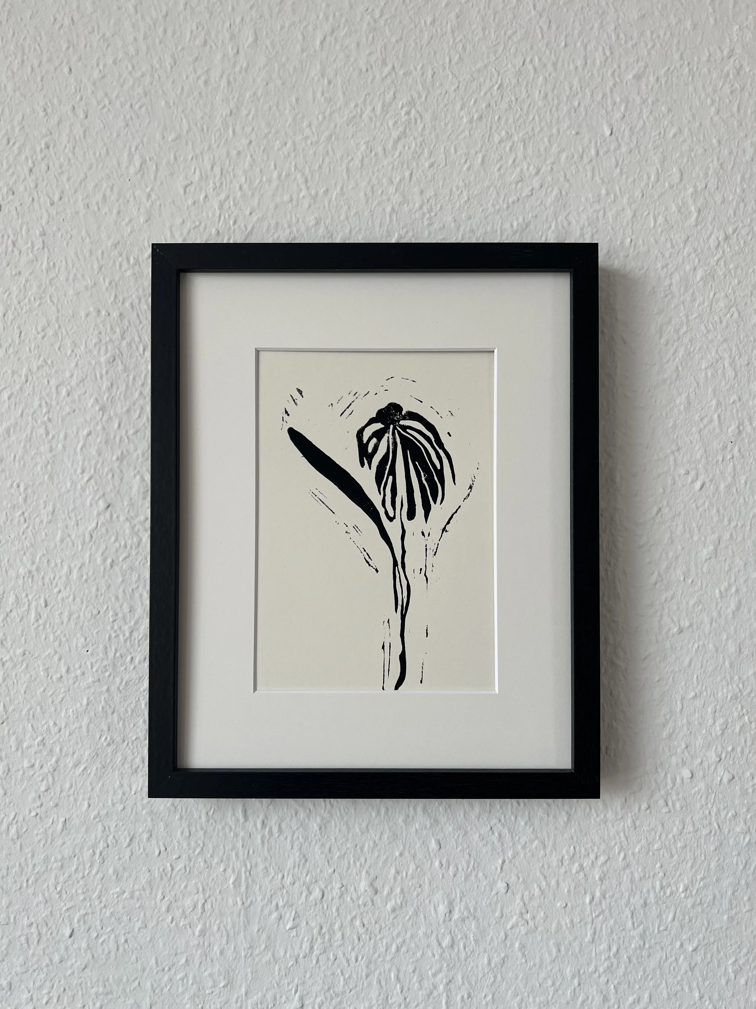 Black and white ink drawing of a flower with delicate, elongated petals, framed and hanging on a textured white wall.