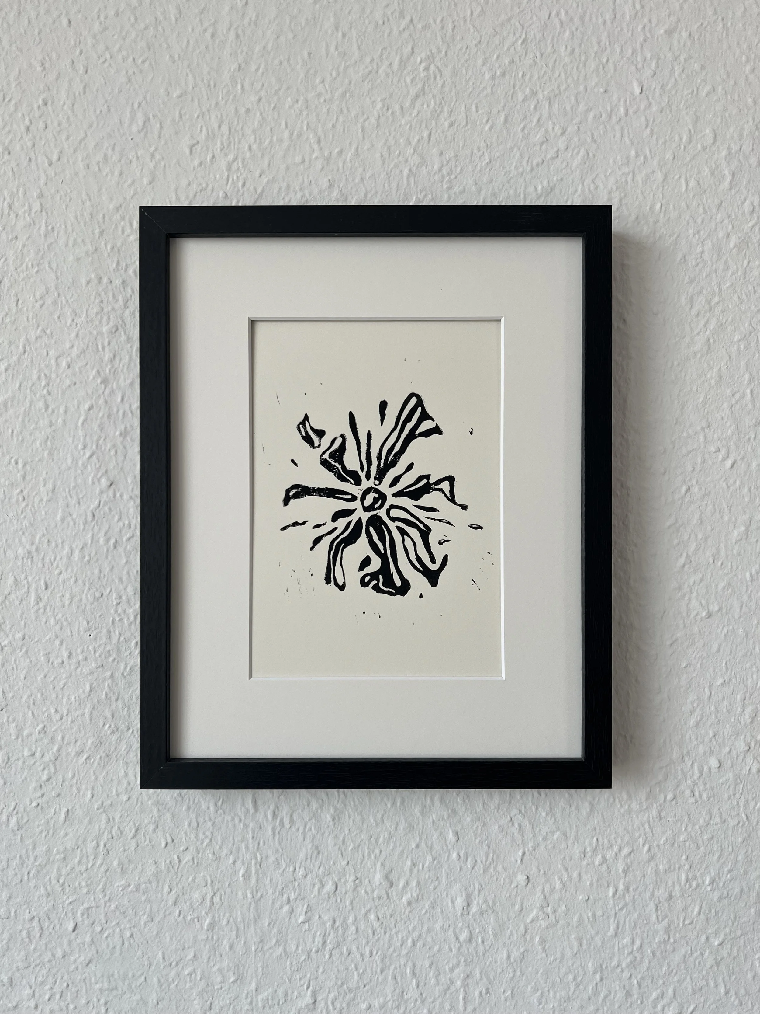 Black frame containing a white mat with a black line drawing of a flower on white paper, hanging on a white textured wall.