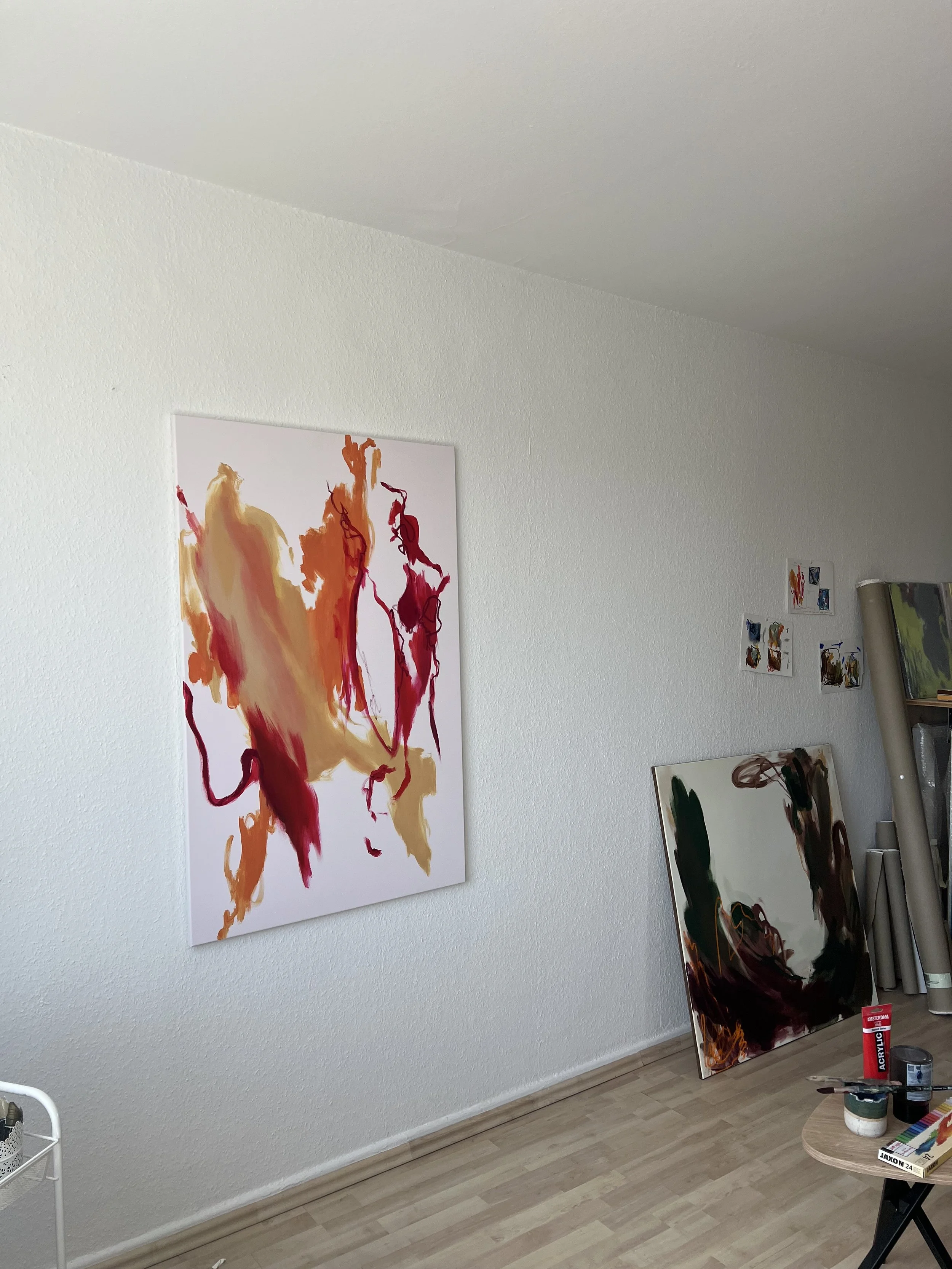 An art gallery displaying abstract paintings on white walls, with one large colorful painting depicting fiery red, orange, and beige tones hanging on the left wall and several smaller artworks on the right wall. A table in the foreground holds painti