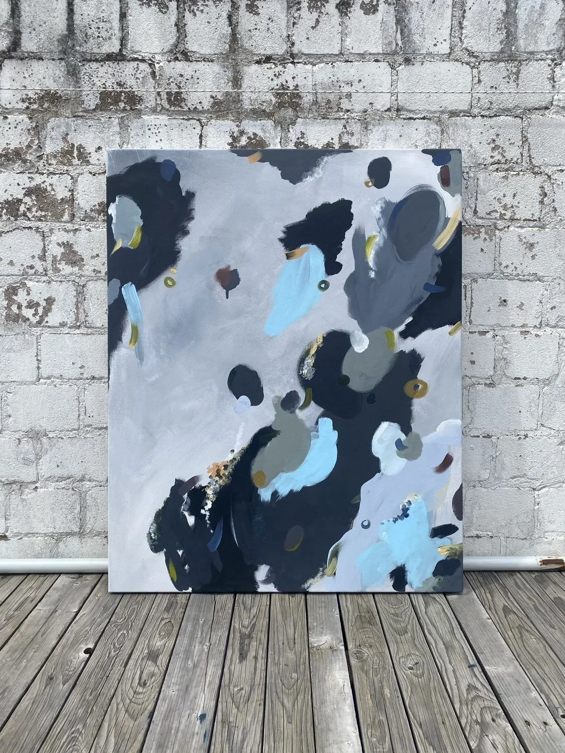 Abstract painting with black, gray, white, blue, brown, and gold brush strokes leaning against a exposed brick wall on a wooden floor.